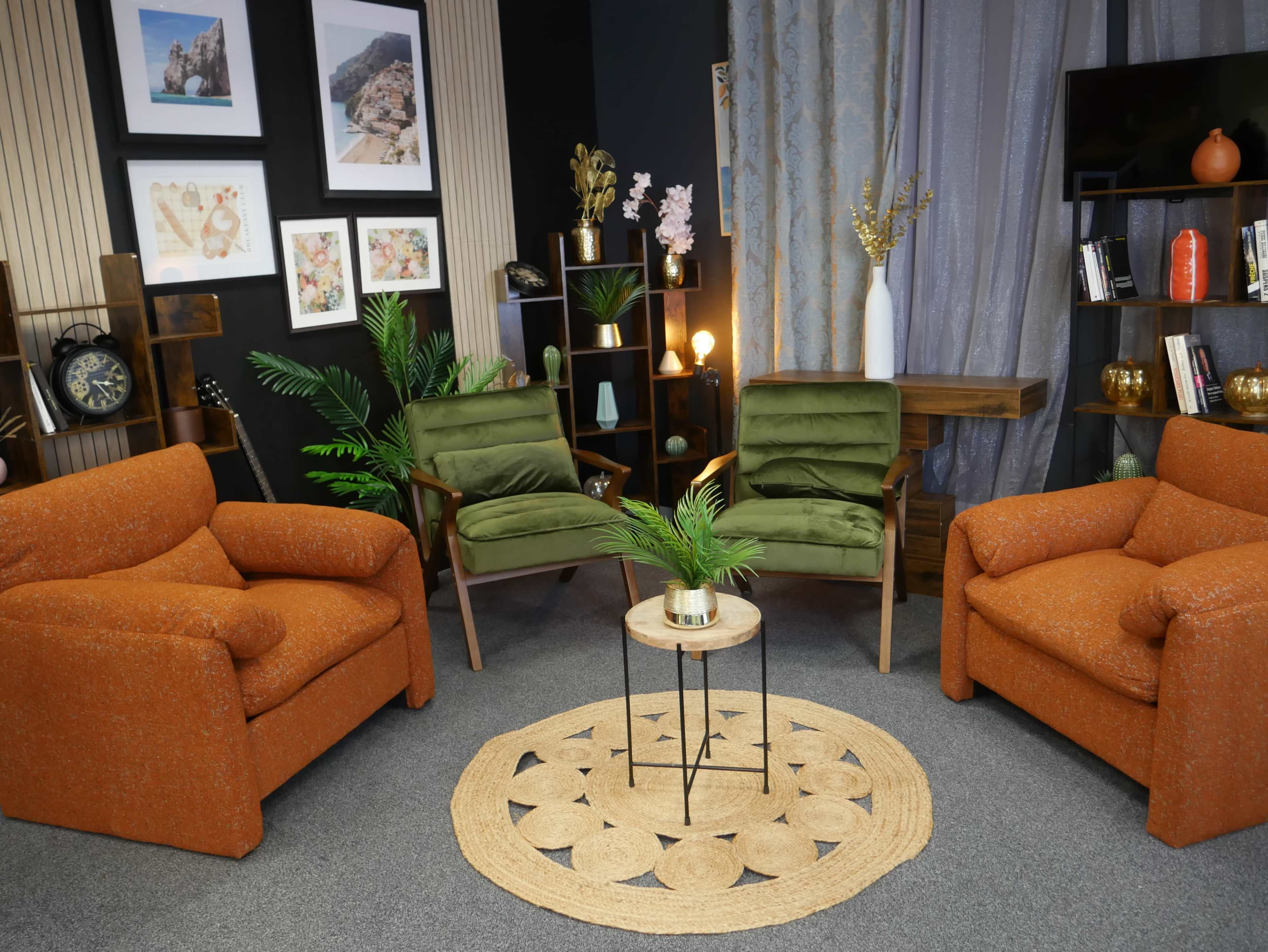 The image shows a cozy interior setting with two orange armchairs, two green chairs, a small round table, and decorative plants, surrounded by artwork and shelves.