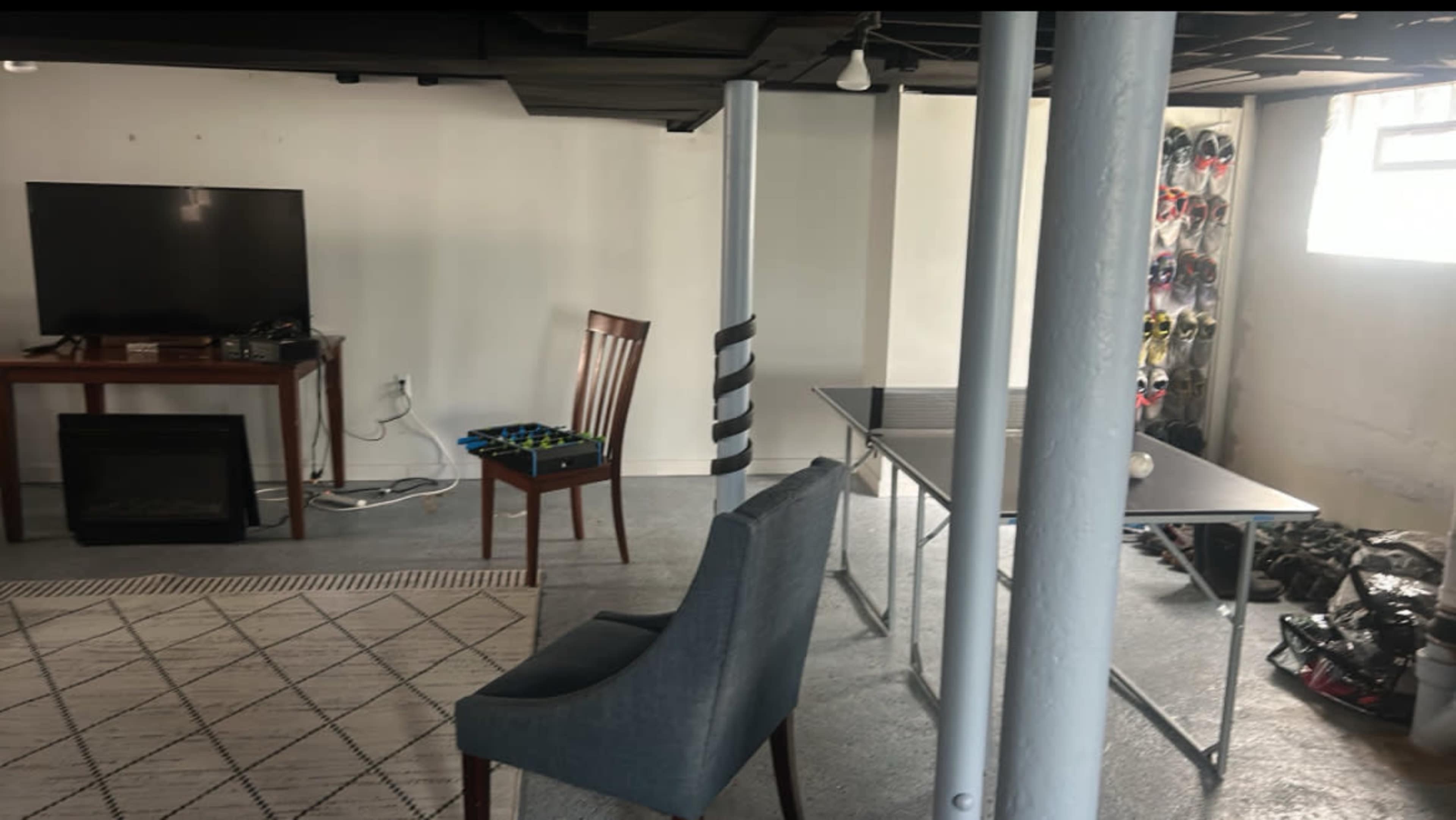 The image shows a basement area with a television on a wooden stand, a chair, a folding table, and a wall lined with shoes.
