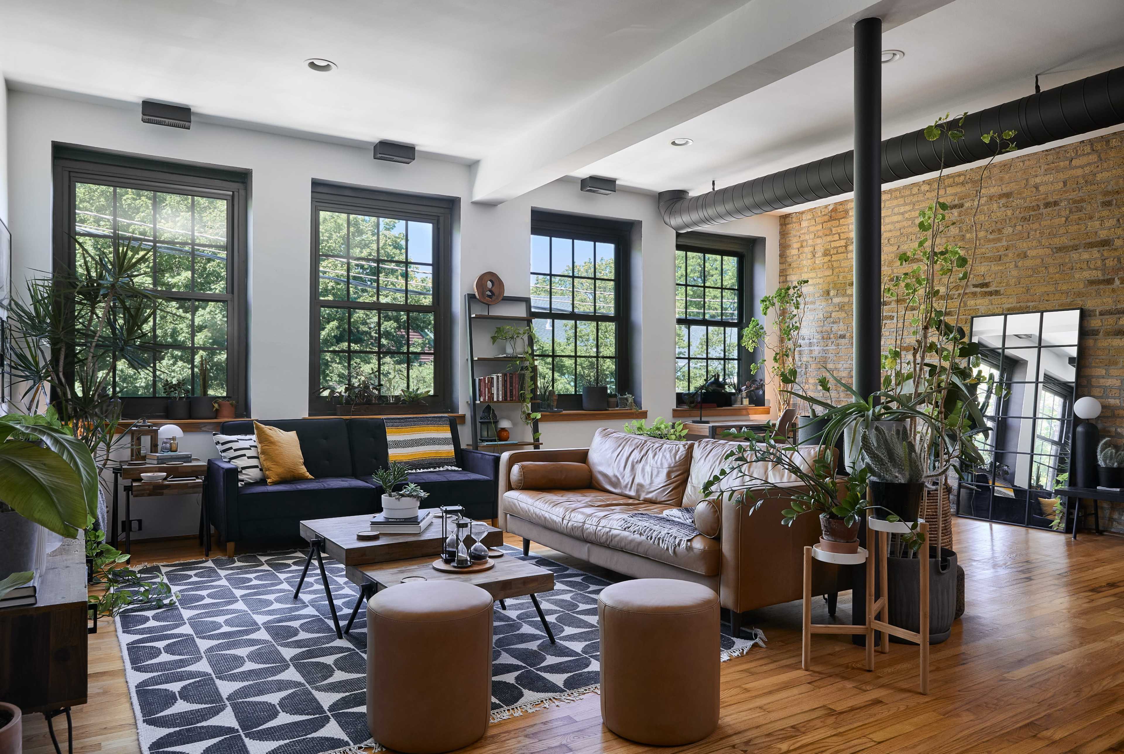 The image depicts a modern living room with large windows, a leather sofa, a coffee table, and various plants, accented by exposed brick walls and a patterned rug.