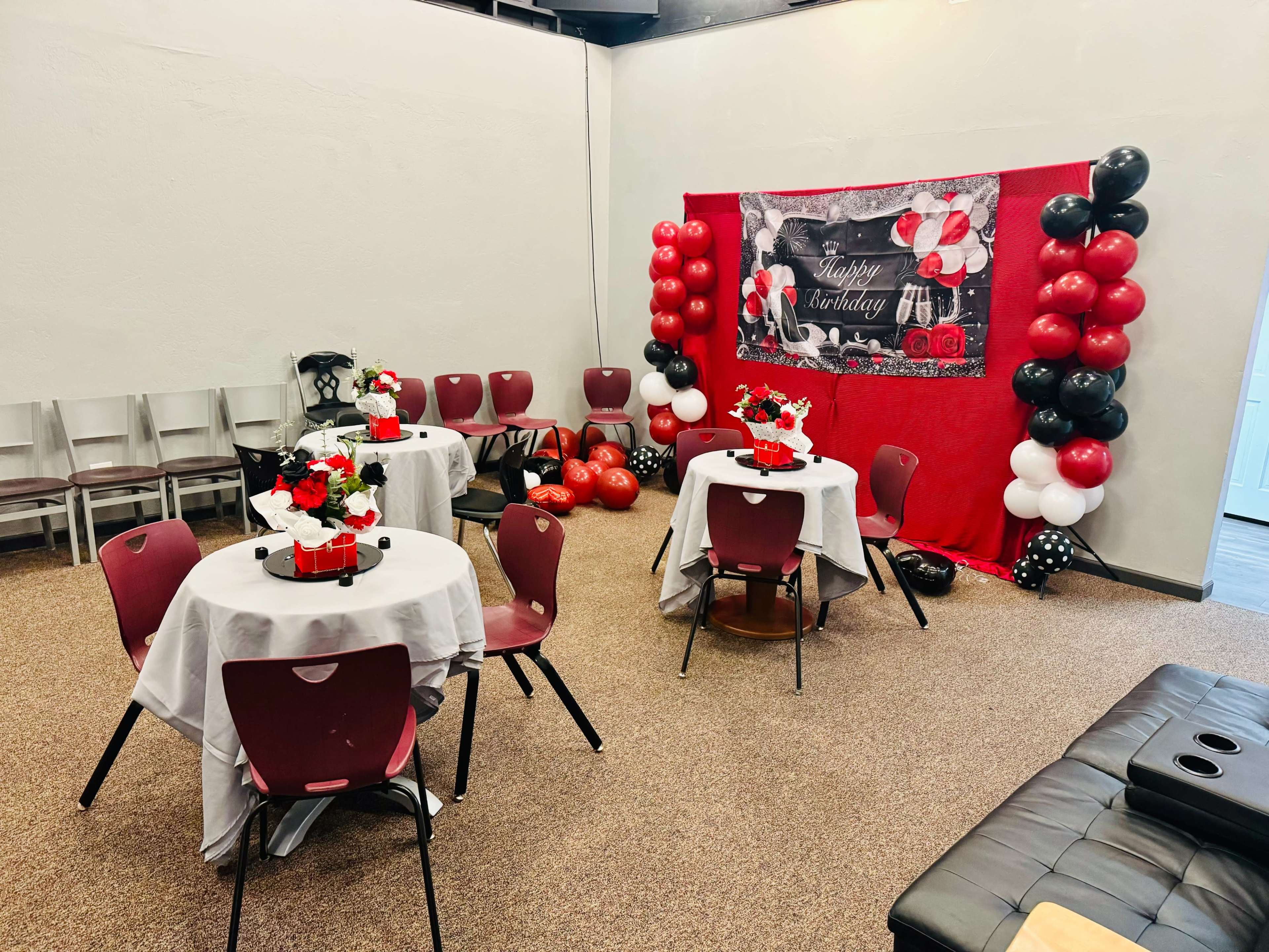 The image shows a decorated party room featuring tables with white tablecloths, red and black balloons, and a birthday banner on the wall.