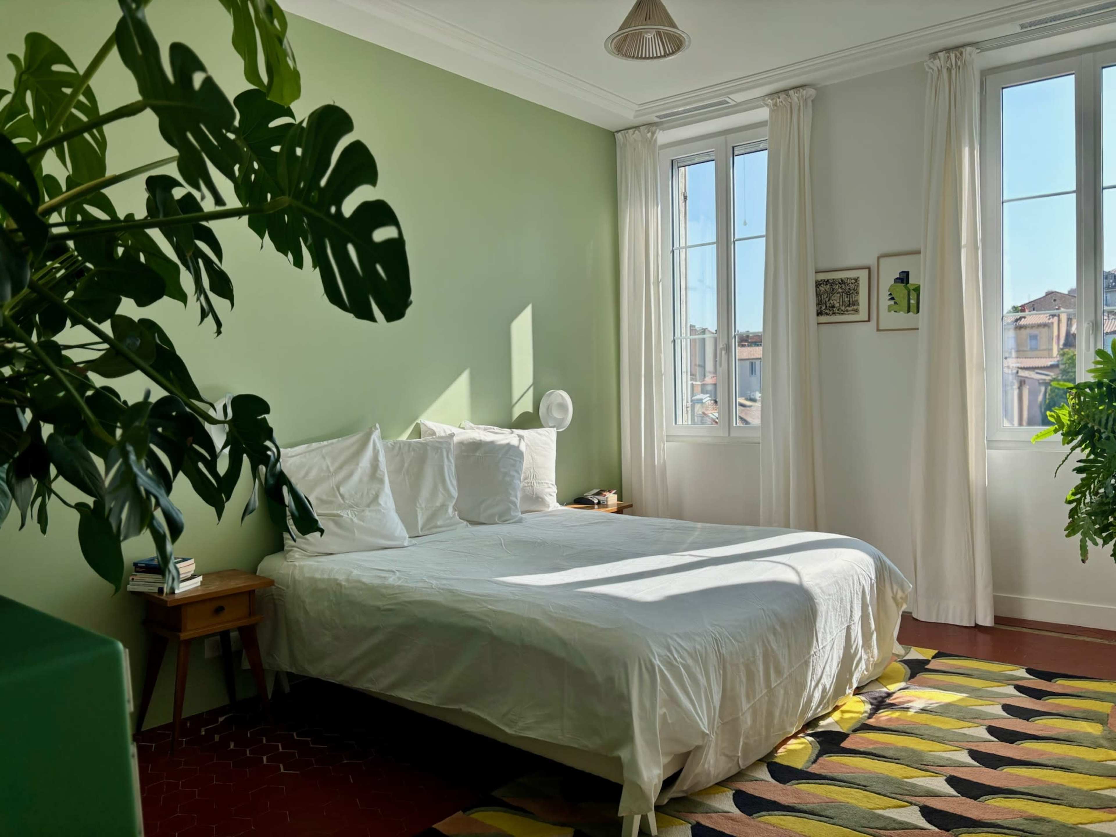 The image shows a bright bedroom with a green accent wall, a white bed with unmade bedding, and large windows allowing sunlight to illuminate the space.