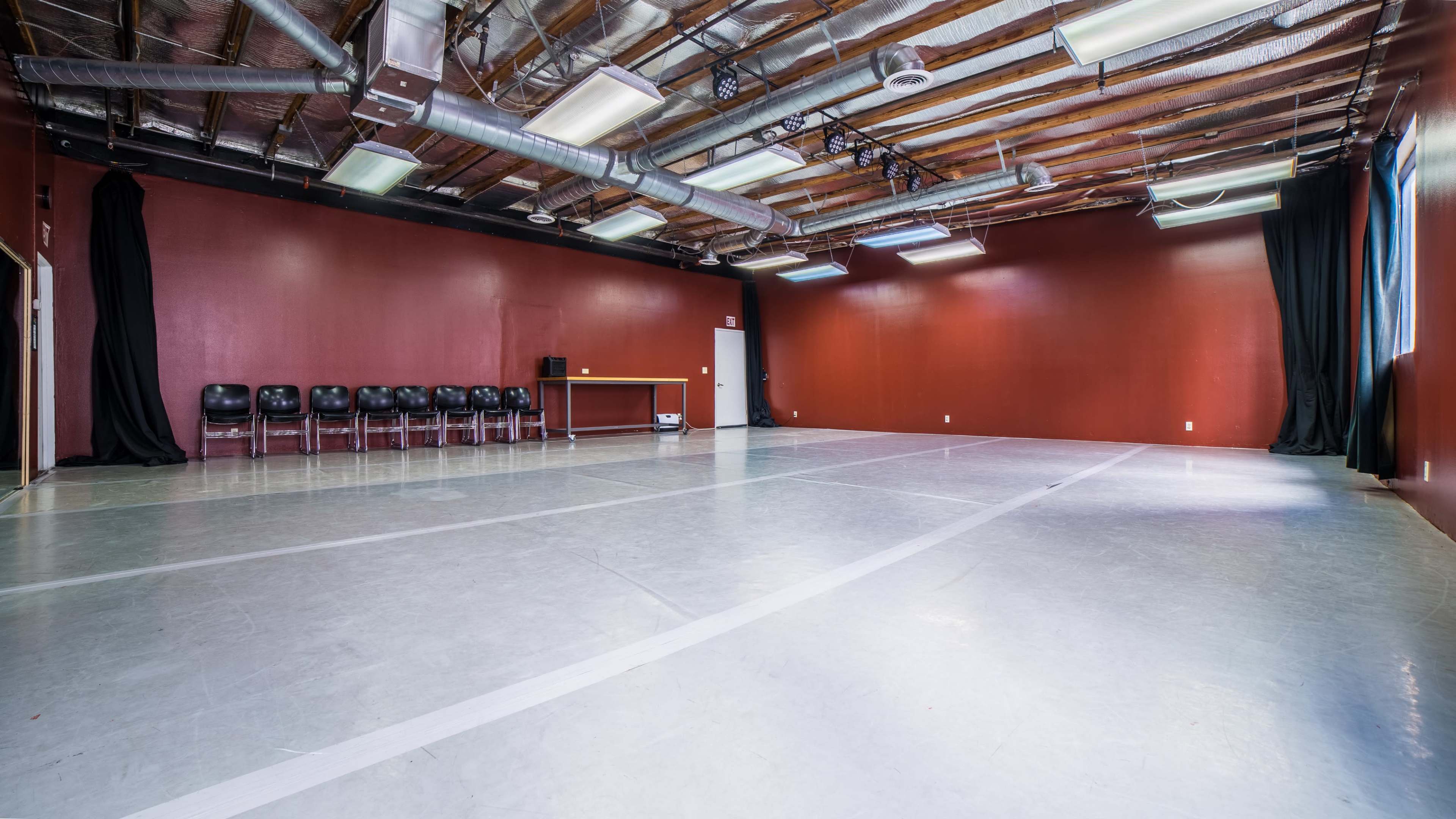 A spacious dance studio with red walls, a gray floor, and a row of black chairs along one side.