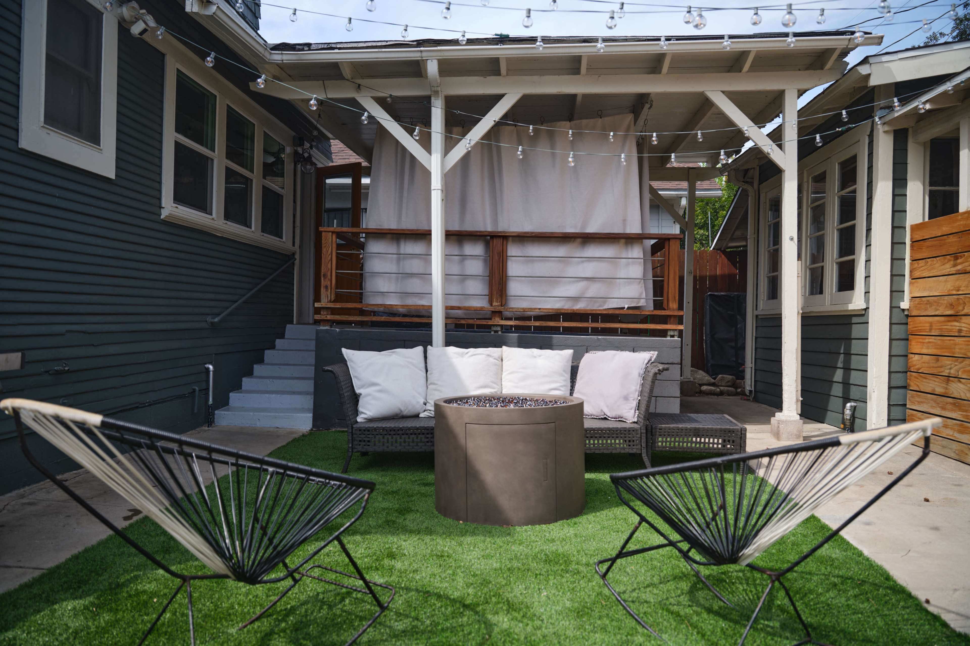 The image shows a backyard patio area with cozy seating, a circular table, and string lights overhead, surrounded by green turf and wooden fencing.