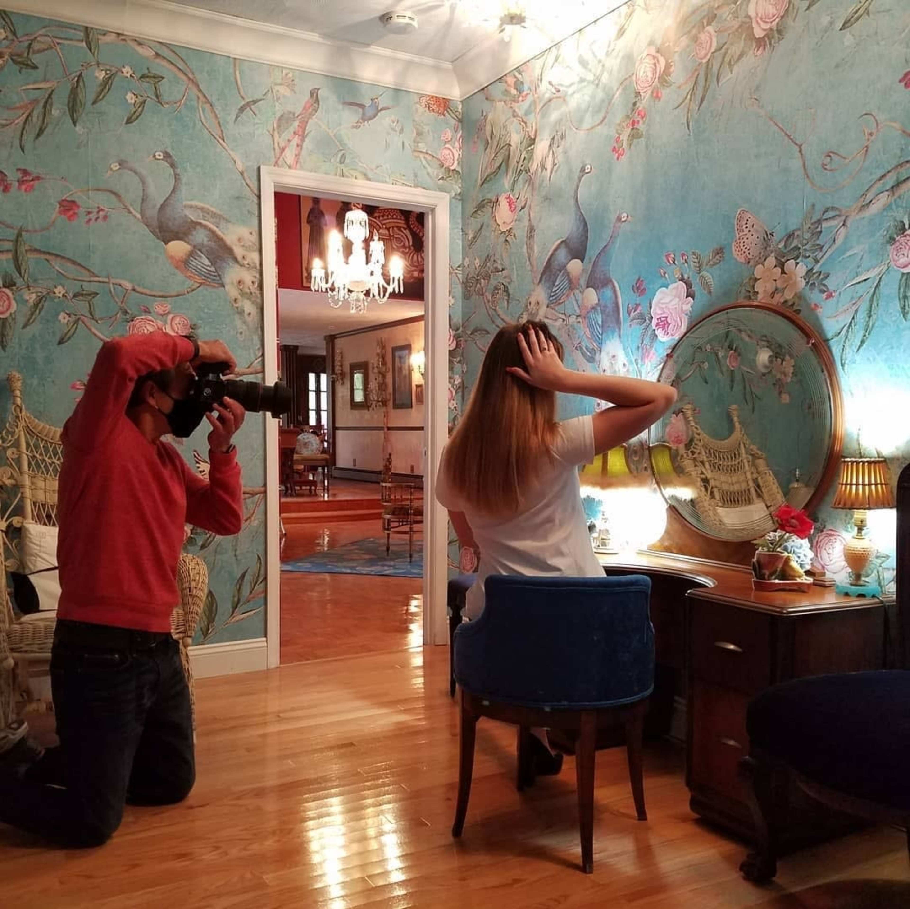 A photographer kneels on the floor while taking a picture of a woman sitting at a vanity in a room with floral wallpaper and decorative lighting.