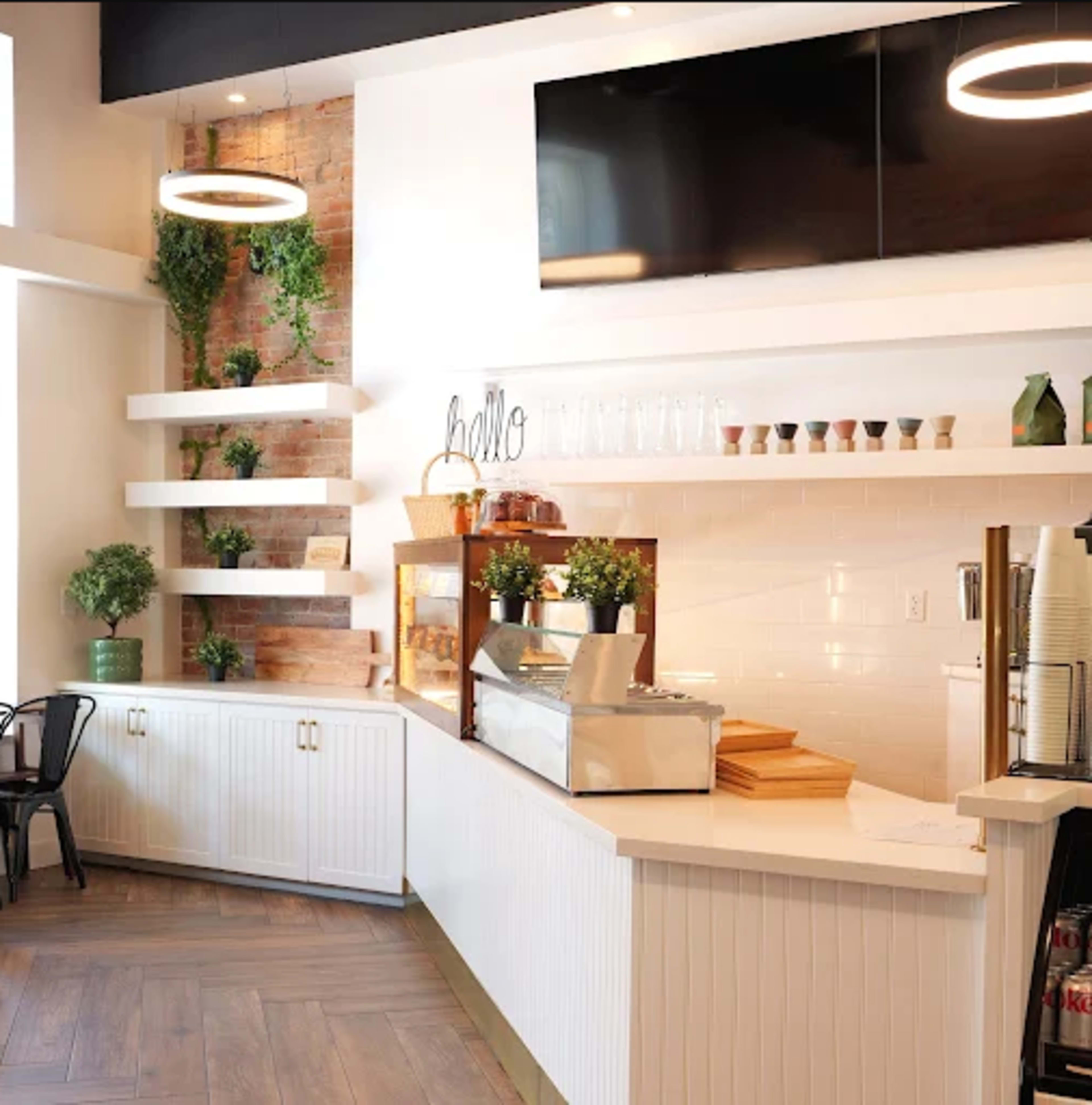 The image shows a modern café interior with a service counter, shelves adorned with plants, and a television mounted on the wall.