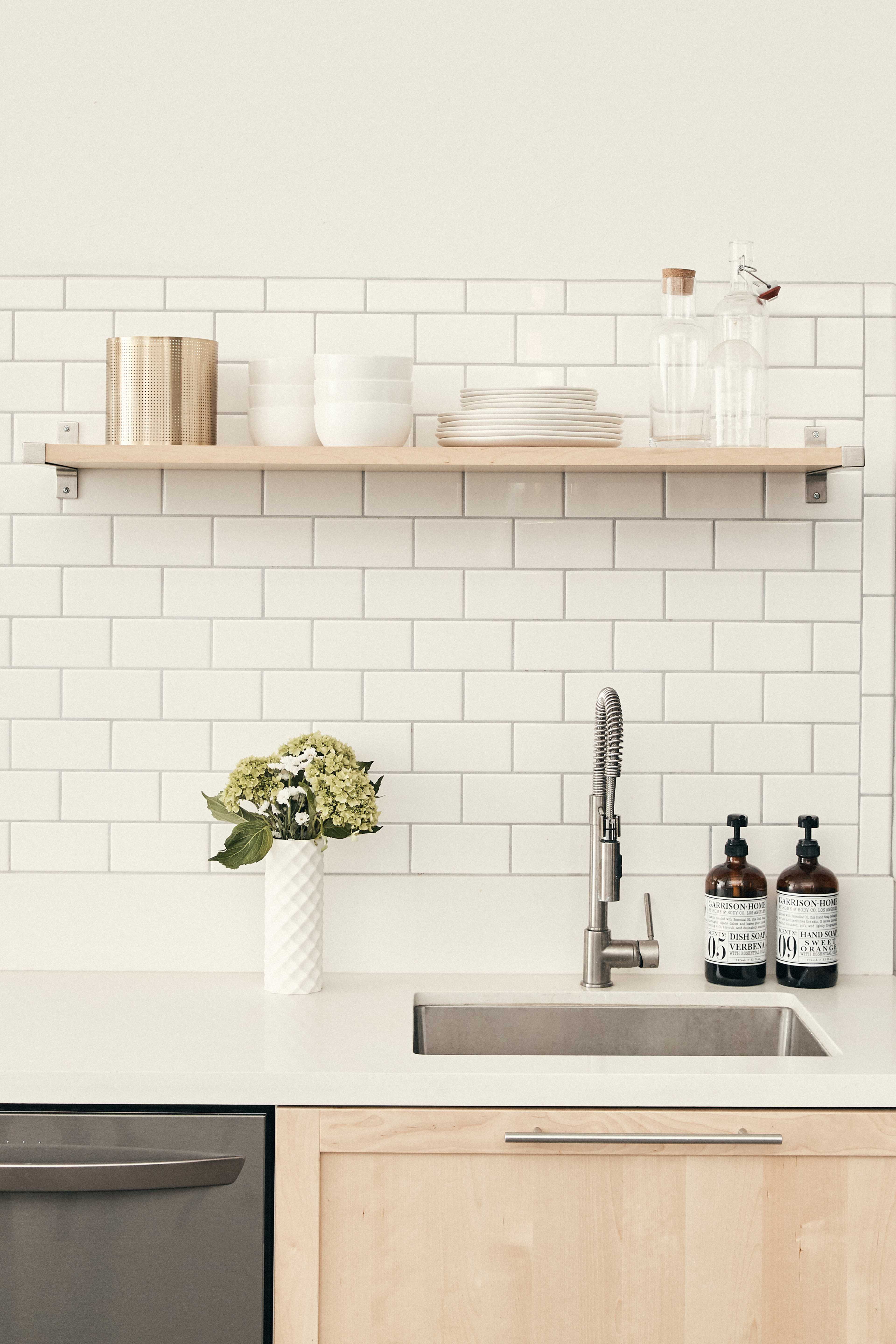 A minimalist kitchen countertop with a sink, a wooden shelf holding dishware, and a vase of flowers.