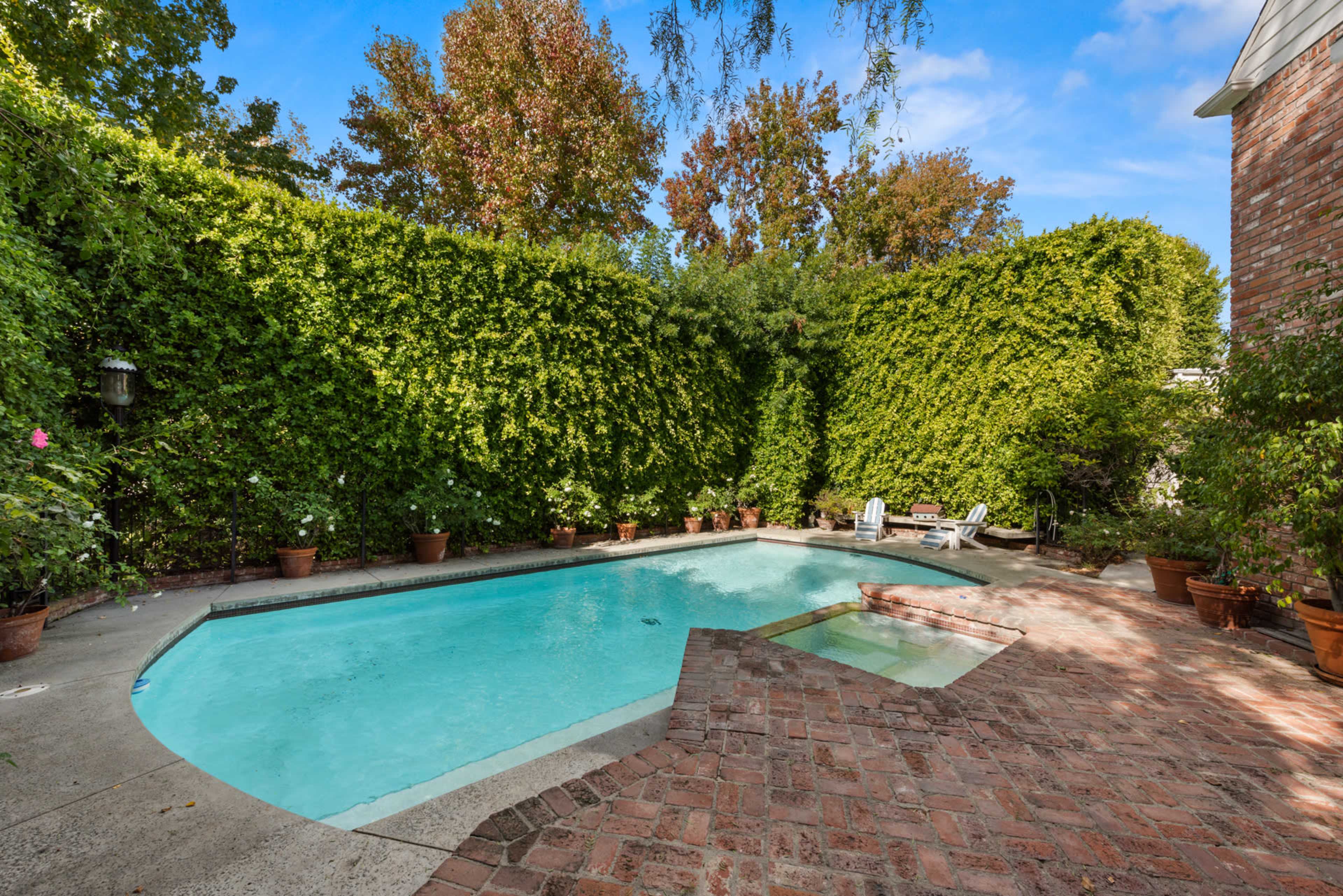 A rectangular swimming pool is surrounded by lush green hedges and potted plants, with a brick patio on one side.