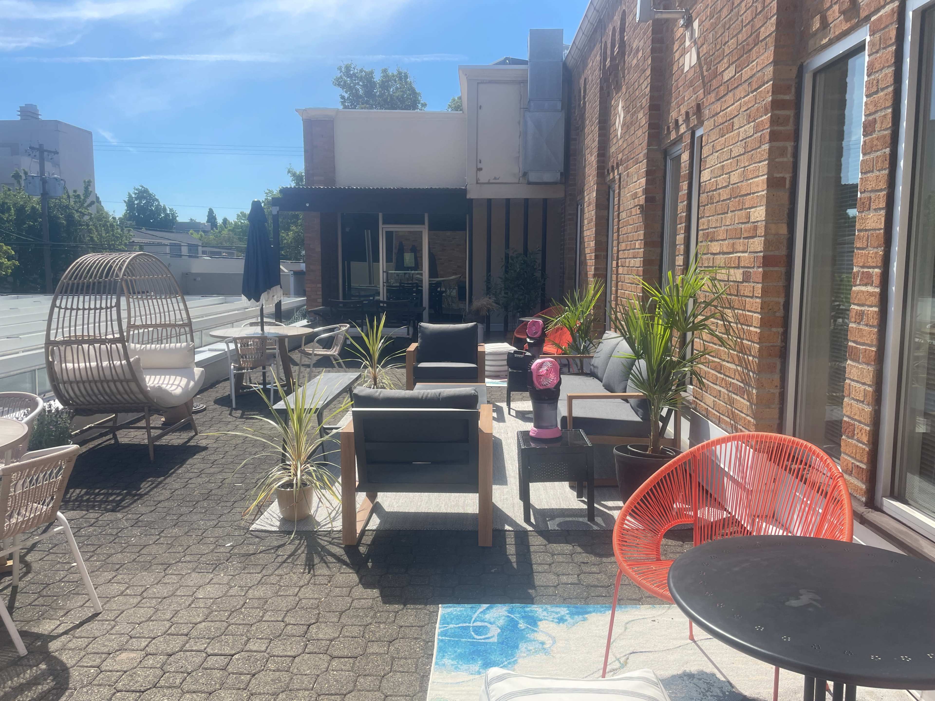 The image shows a patio area with modern seating, potted plants, and a mix of furniture, all under bright sunlight.