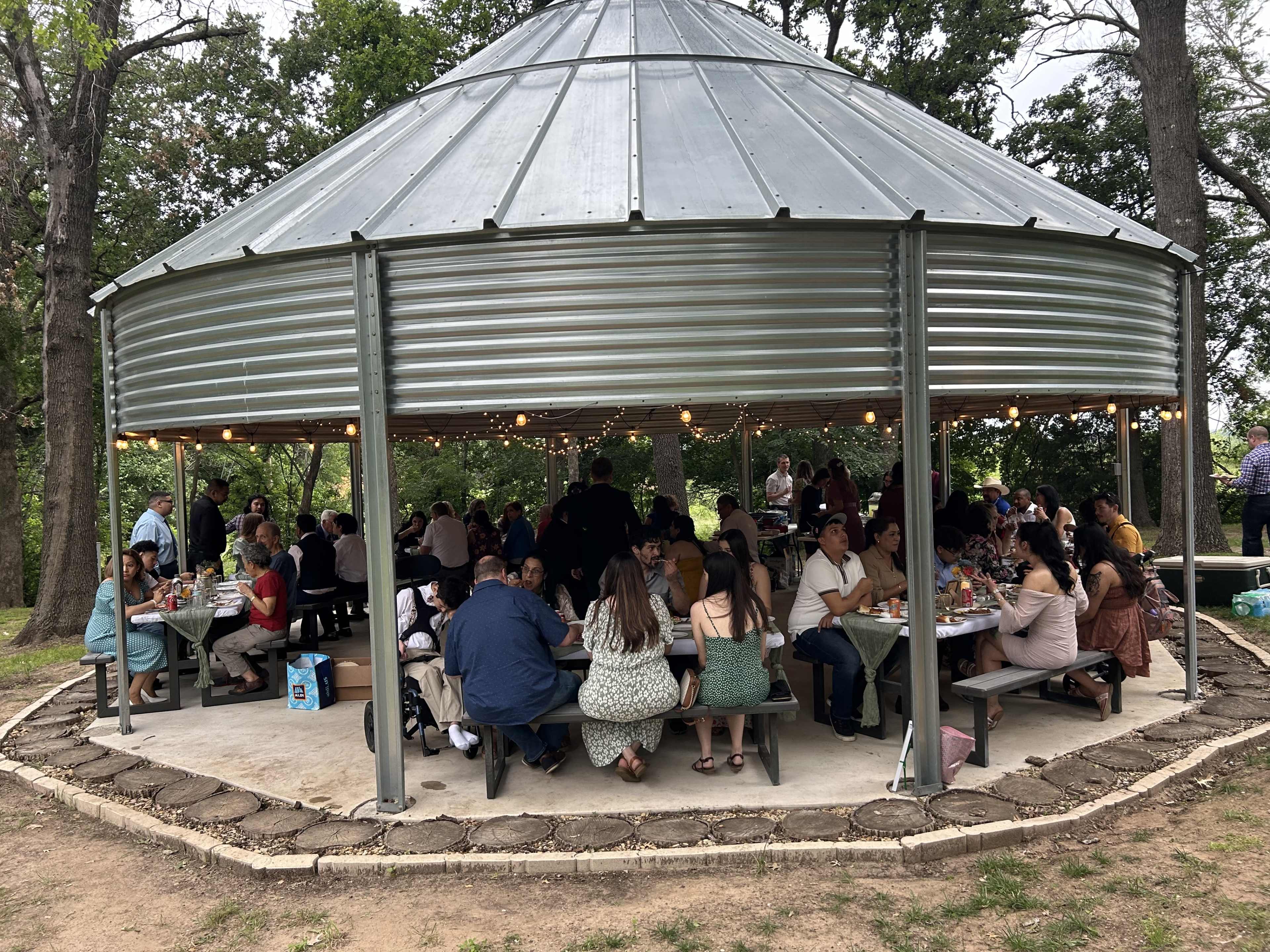 A large group of people is gathered under a circular pavilion with picnic tables, enjoying a meal together.