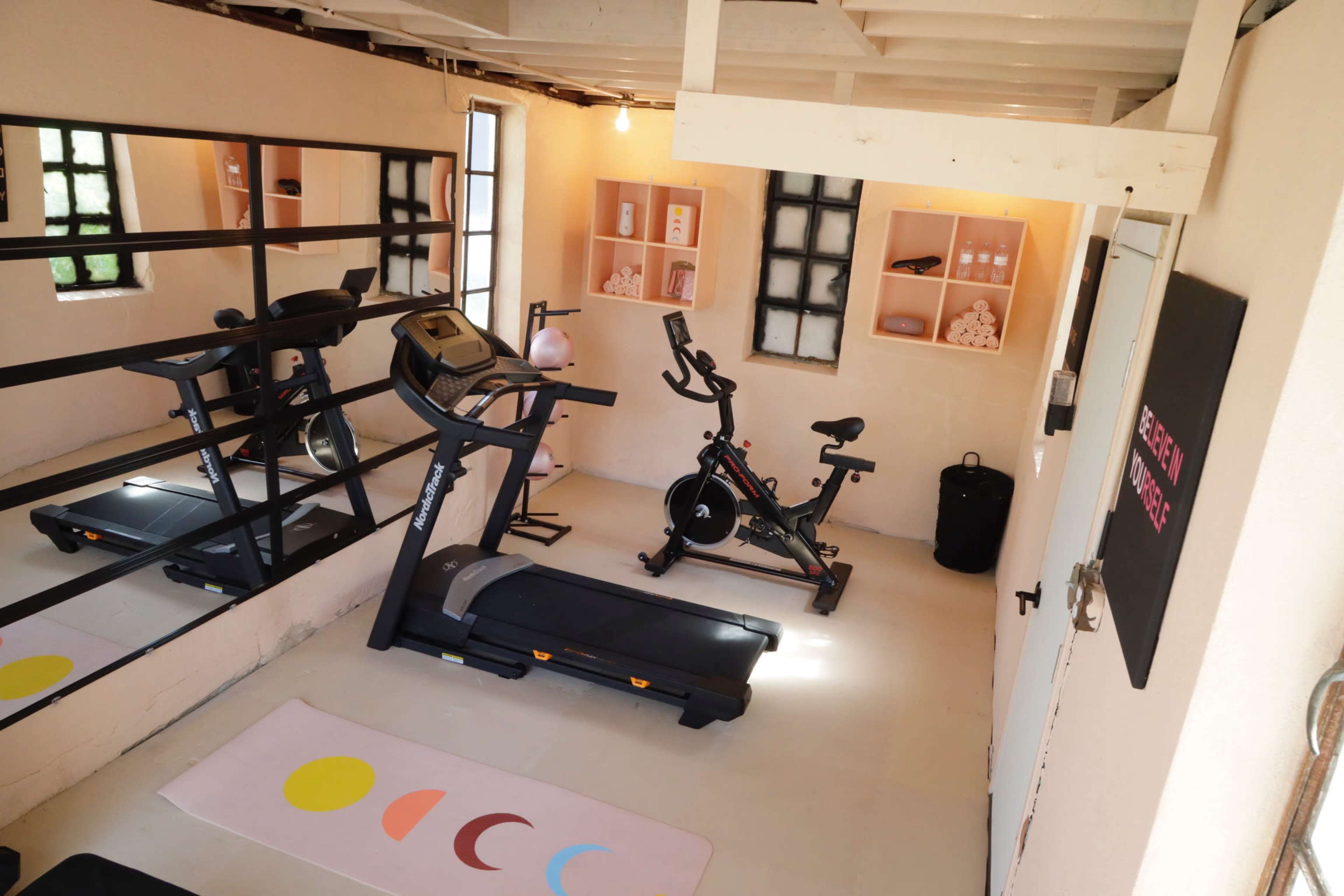 The image shows a small home gym featuring a treadmill and an exercise bike, with mirrors on one wall and neatly organized shelves displaying fitness accessories.