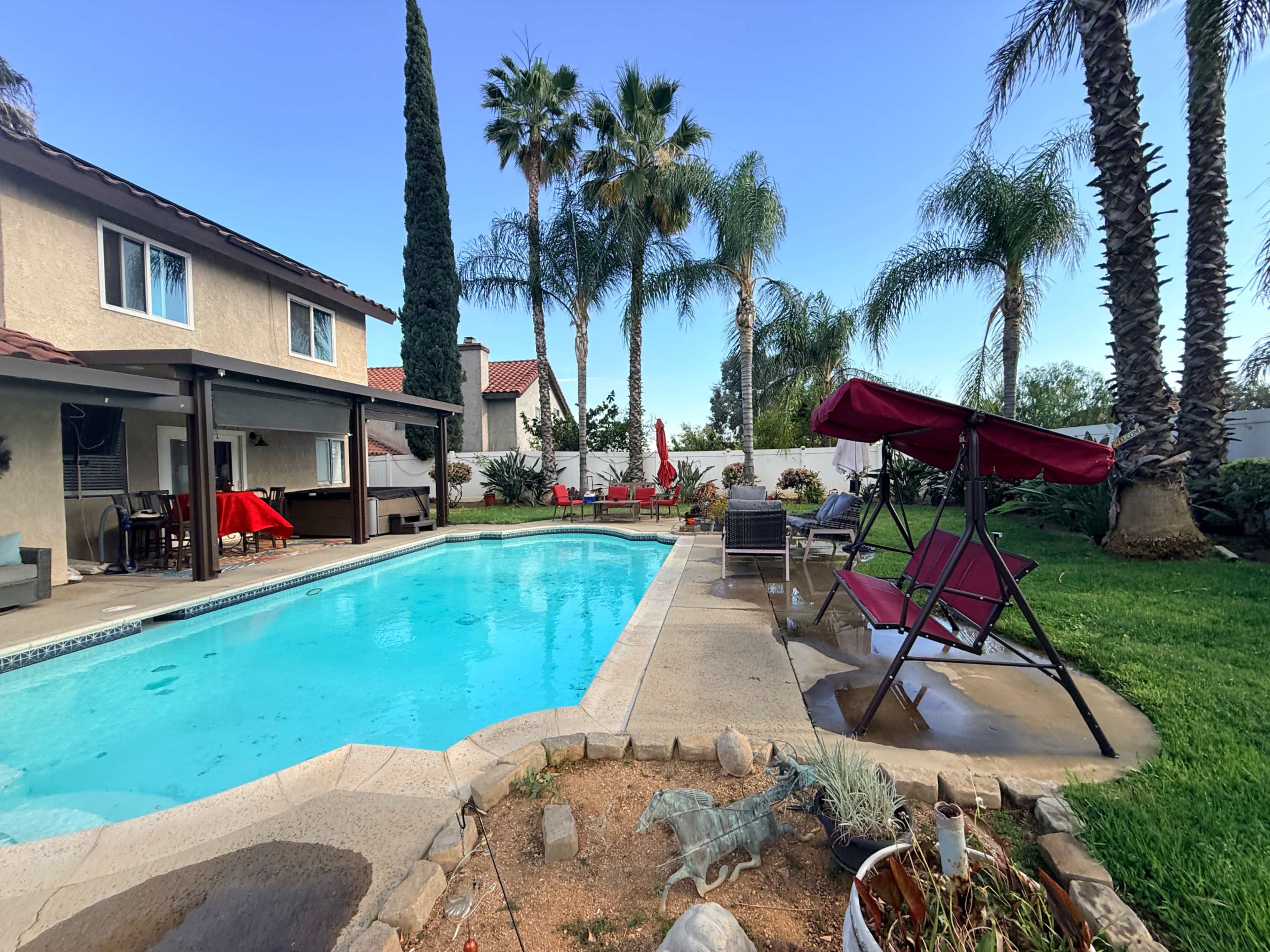 The image shows a backyard with a swimming pool, a shaded seating area, and palm trees in a sunny residential setting.