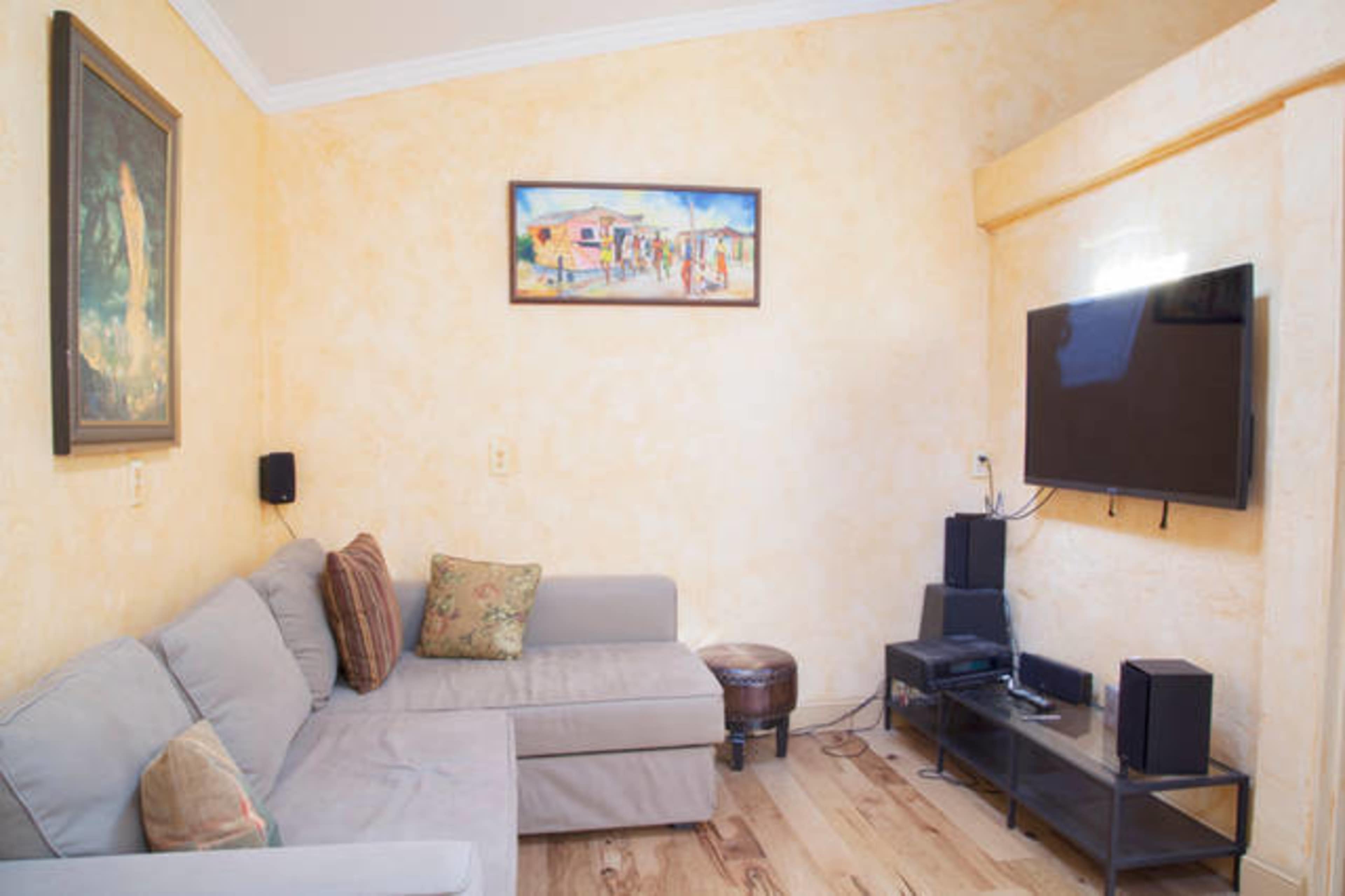 A small living room with a gray sofa, a wooden coffee table, a flat-screen TV mounted on the wall, and a painting hanging above the sofa.