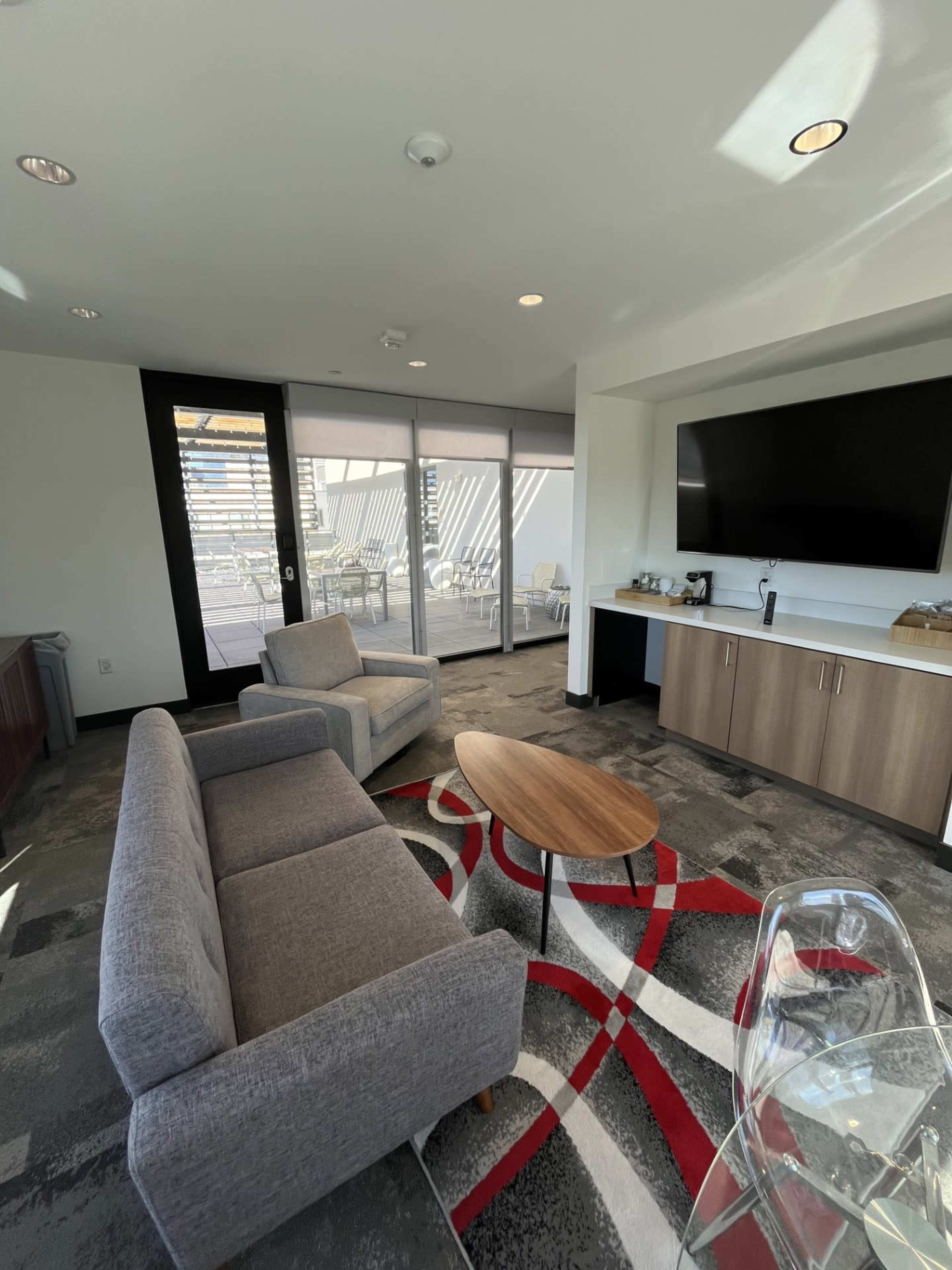 The image shows a modern lounge area featuring two gray sofas, a wooden coffee table, and a television mounted on the wall, with large windows providing natural light.