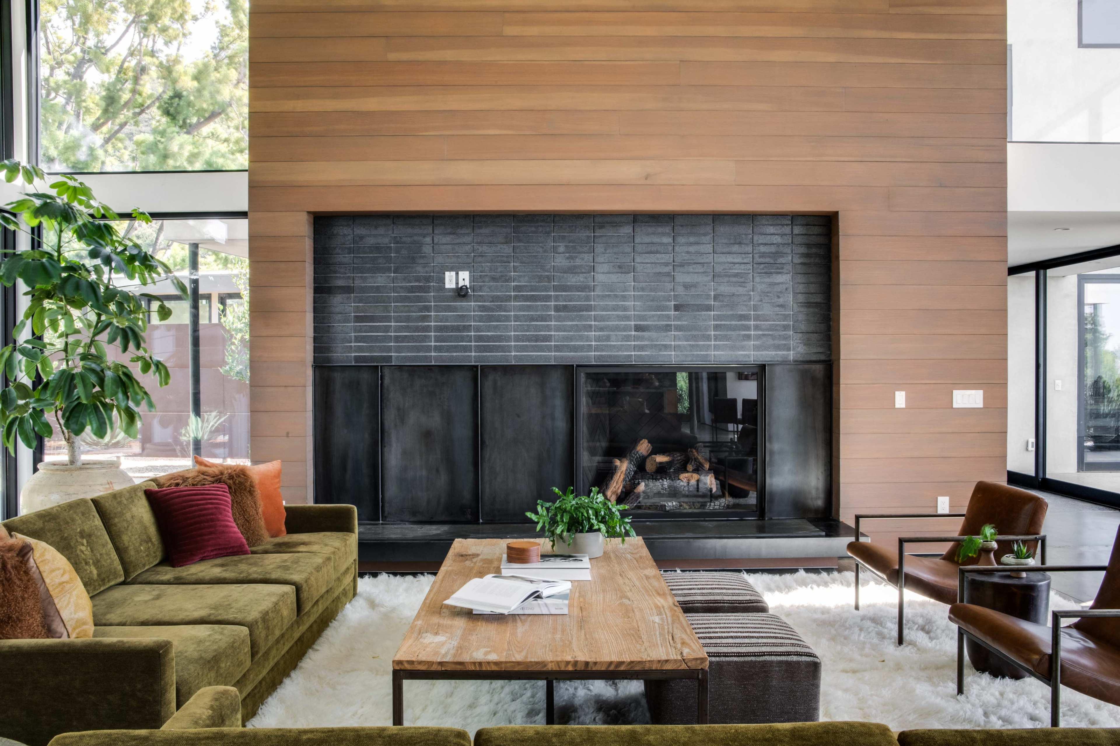 The image shows a modern living room with a large wooden coffee table, a green sofa, an armchair, and a fireplace framed by wooden paneling.