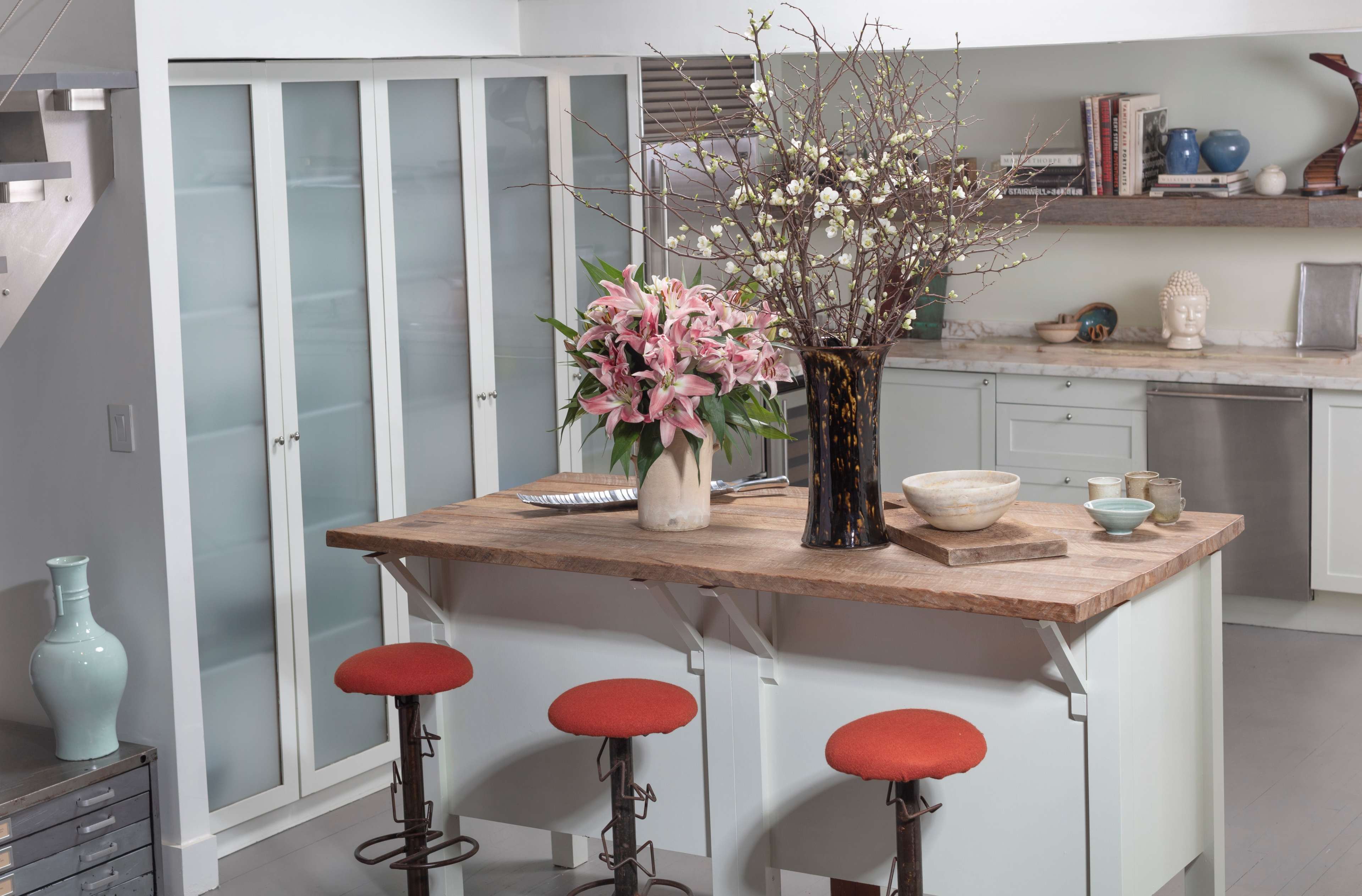 A modern kitchen features a wooden island with two red stools, adorned with a vase of flowers and branches, alongside white cabinetry and a marble countertop.
