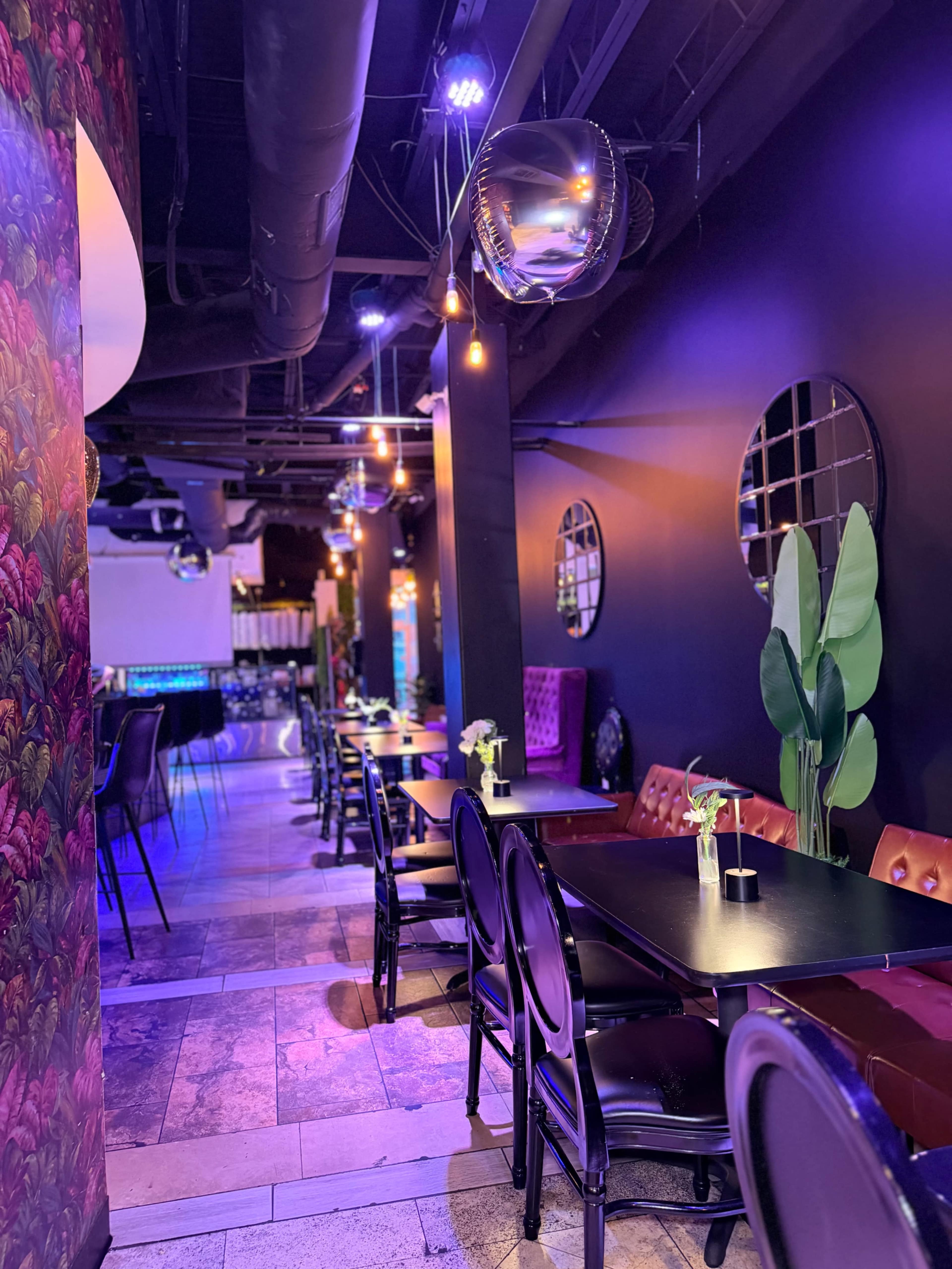 The image shows a modern restaurant interior with black tables and chairs, purple lighting, and decorative plants along the walls.