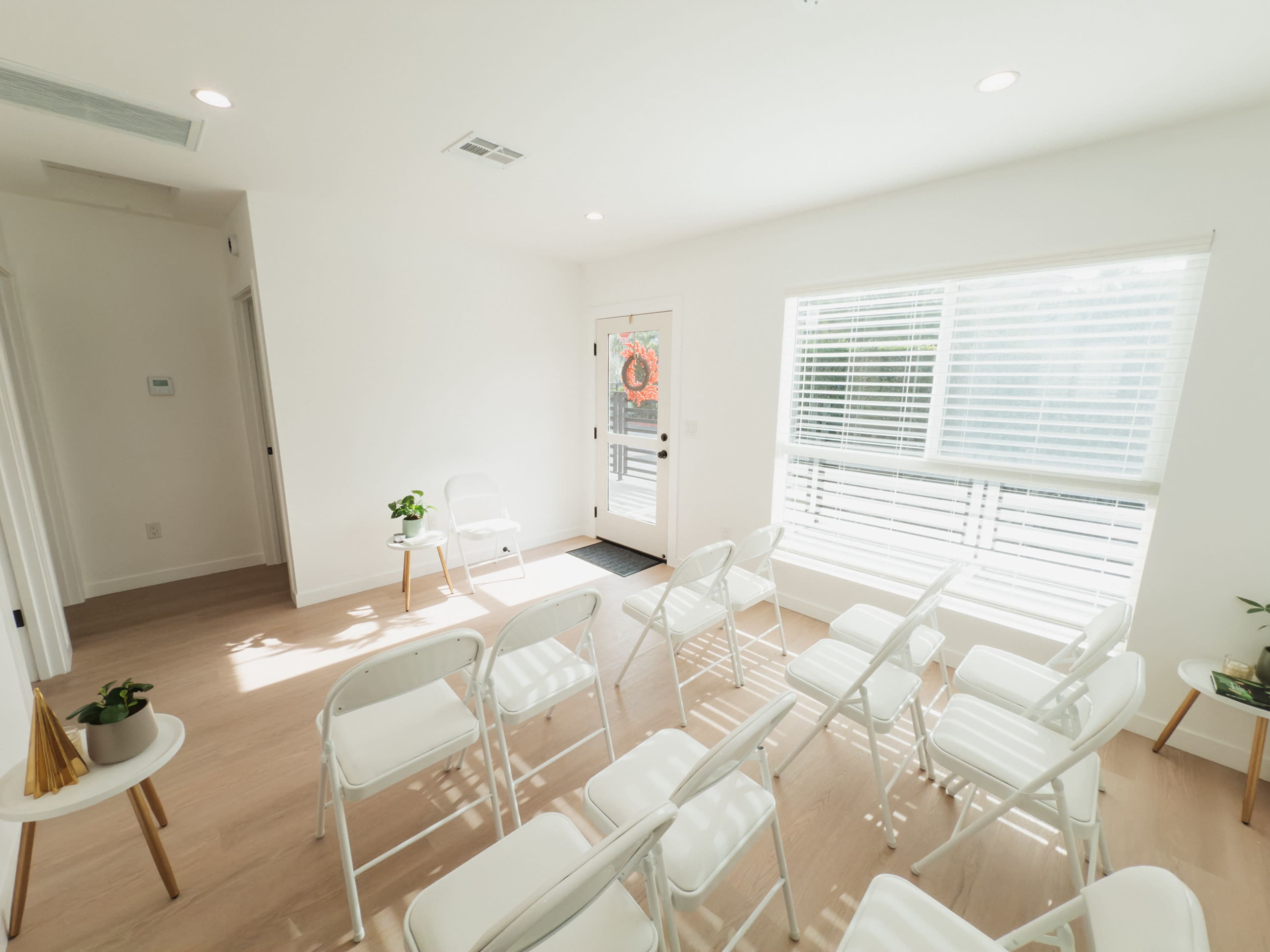 A bright, sparsely furnished waiting room features white folding chairs arranged in rows, with a door and large windows allowing natural light to enter.