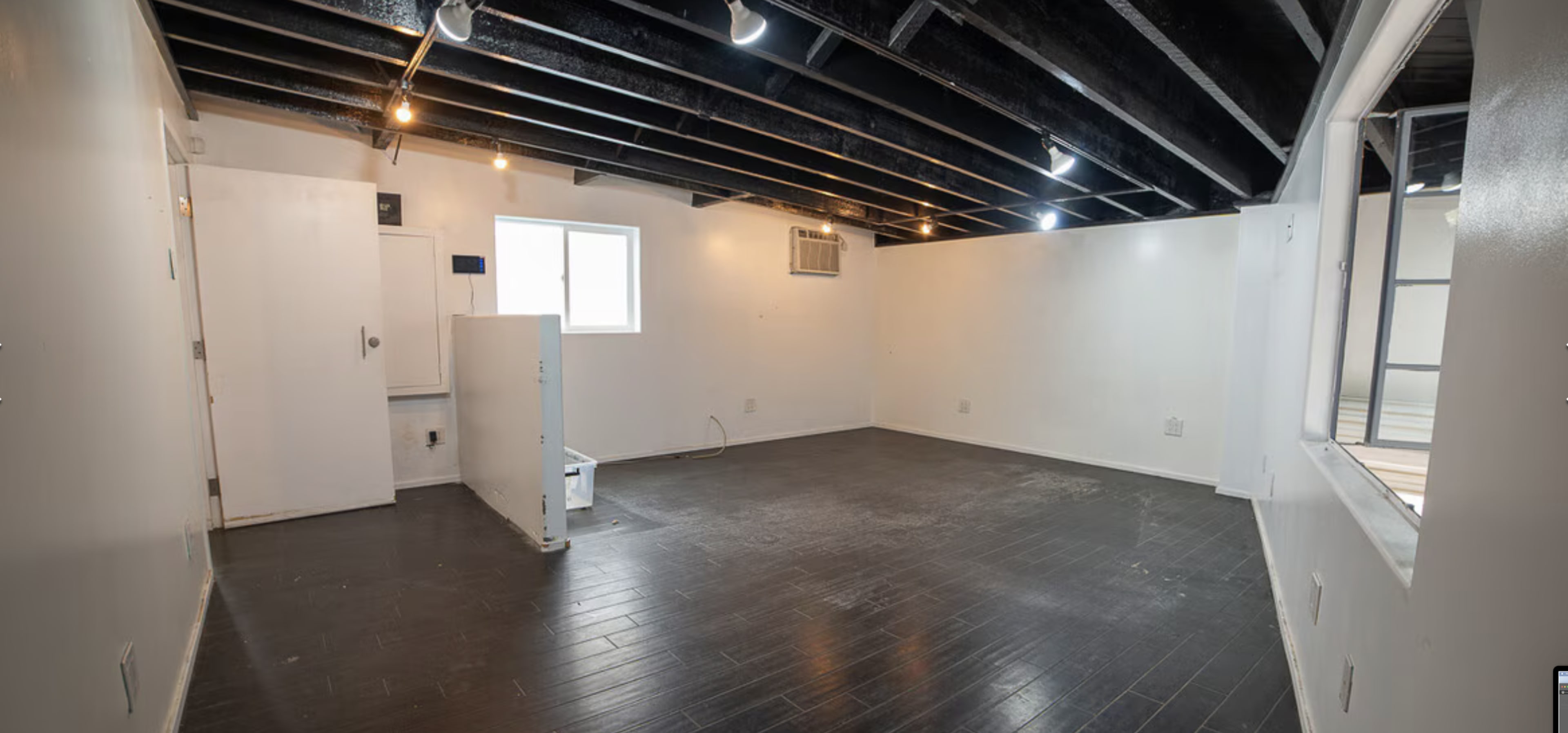 The image shows a sparsely furnished room with black exposed beams, white walls, and dark wood flooring, featuring a window and an air conditioning unit.