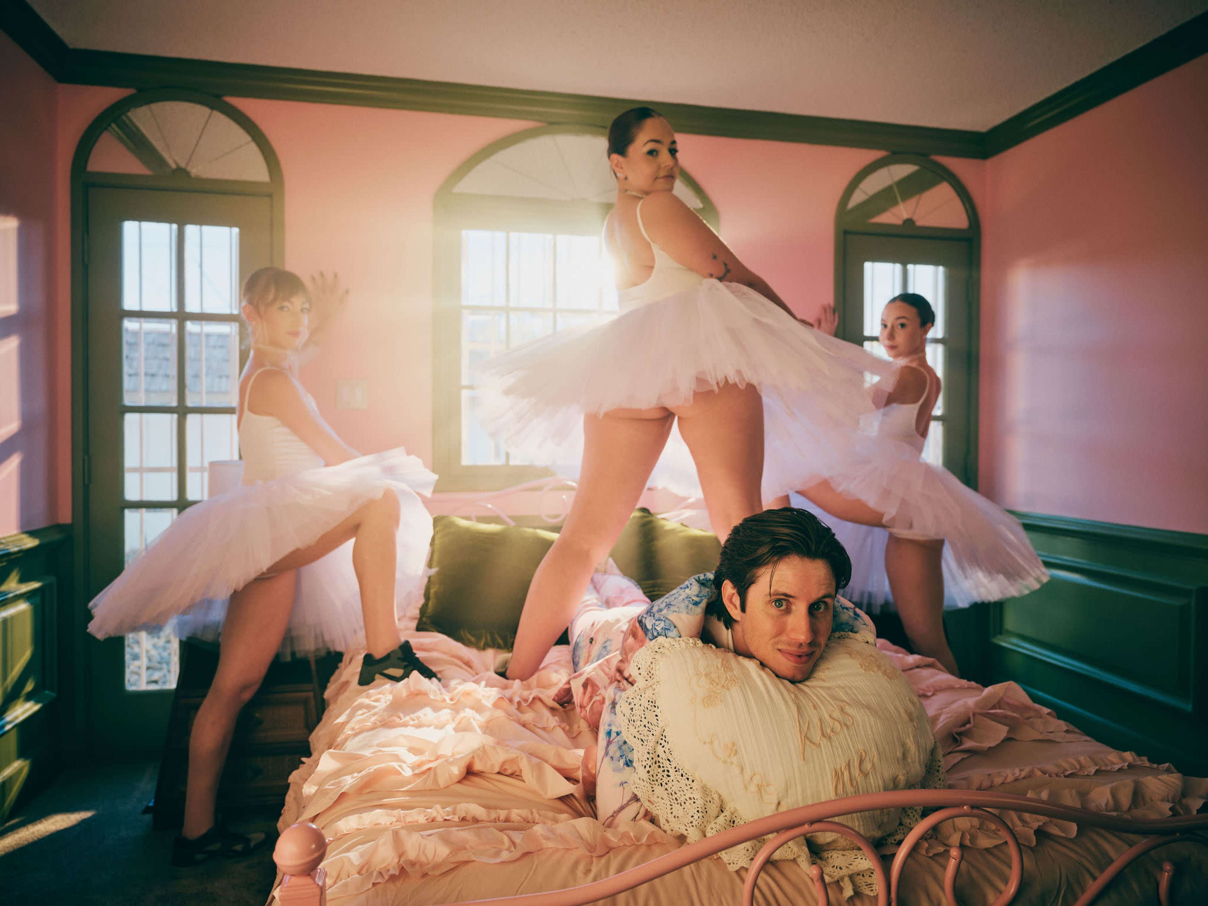 A man reclines on a bed surrounded by three ballet dancers in tutus, set against a pastel pink bedroom.