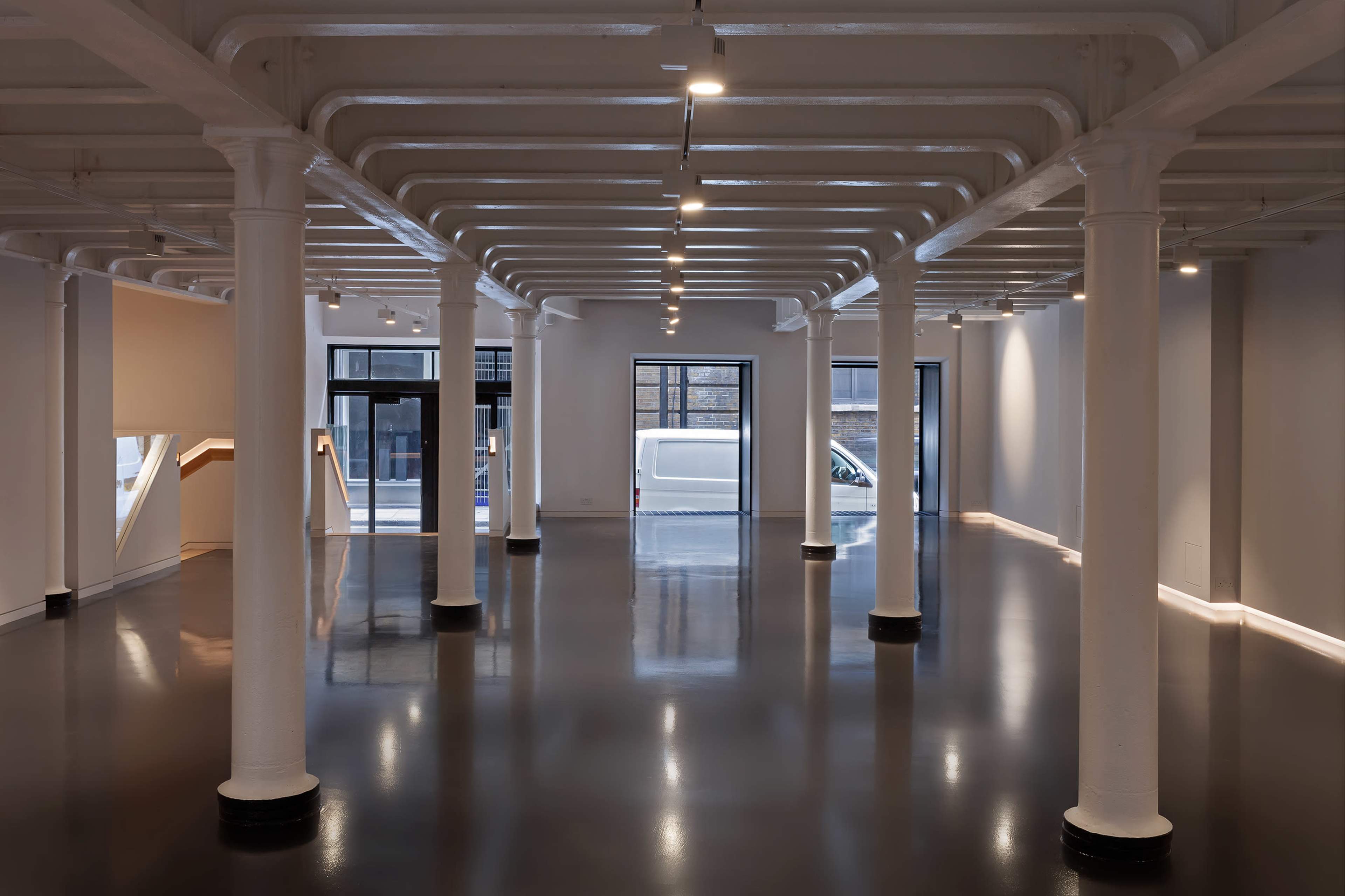 A spacious, vacant interior of an art gallery with white pillars supporting the ceiling and large windows allowing natural light from the street outside.