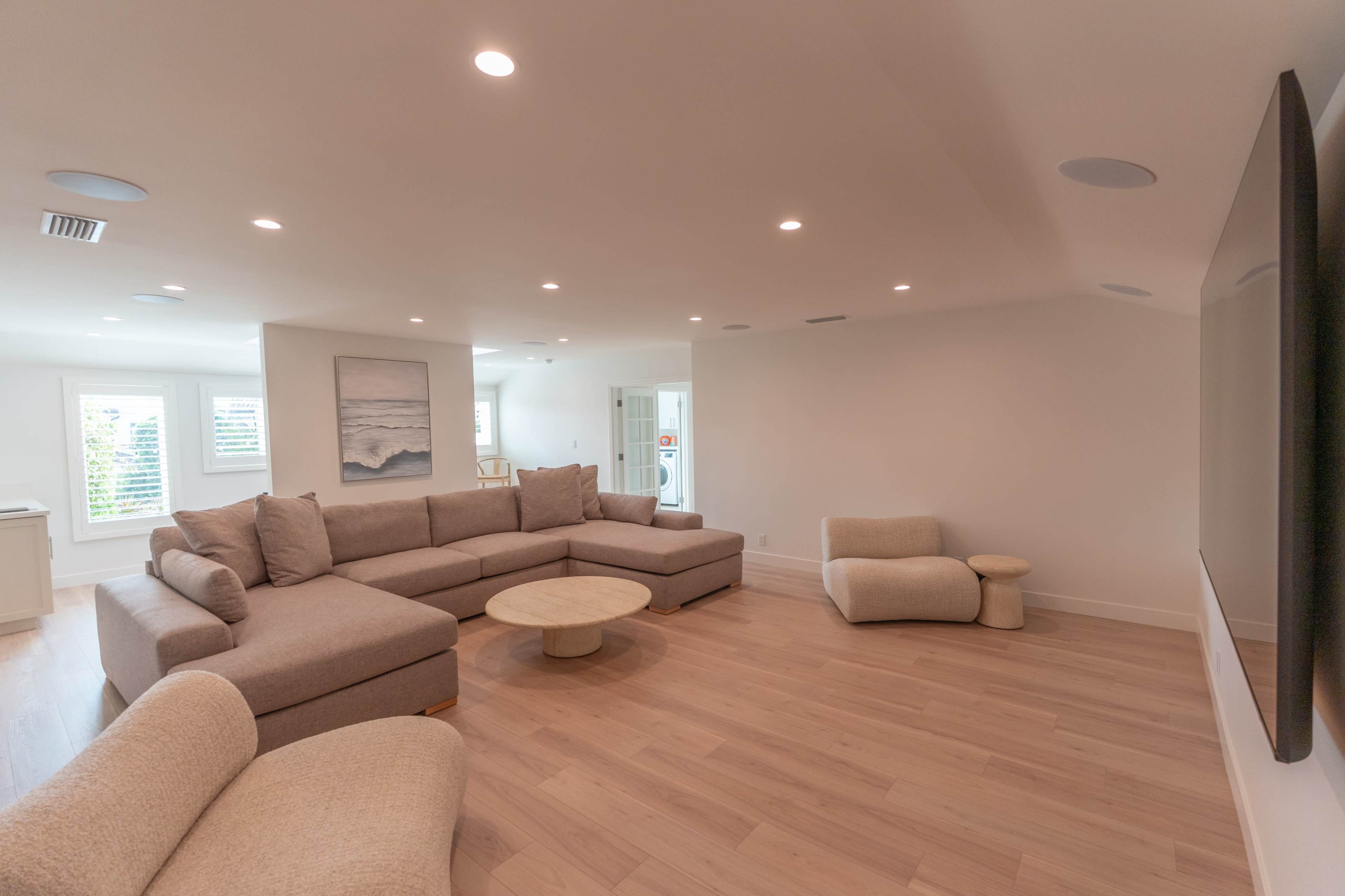 The image shows a modern living room featuring a large sectional sofa, a round coffee table, and a wall-mounted TV in a bright, minimalist setting.