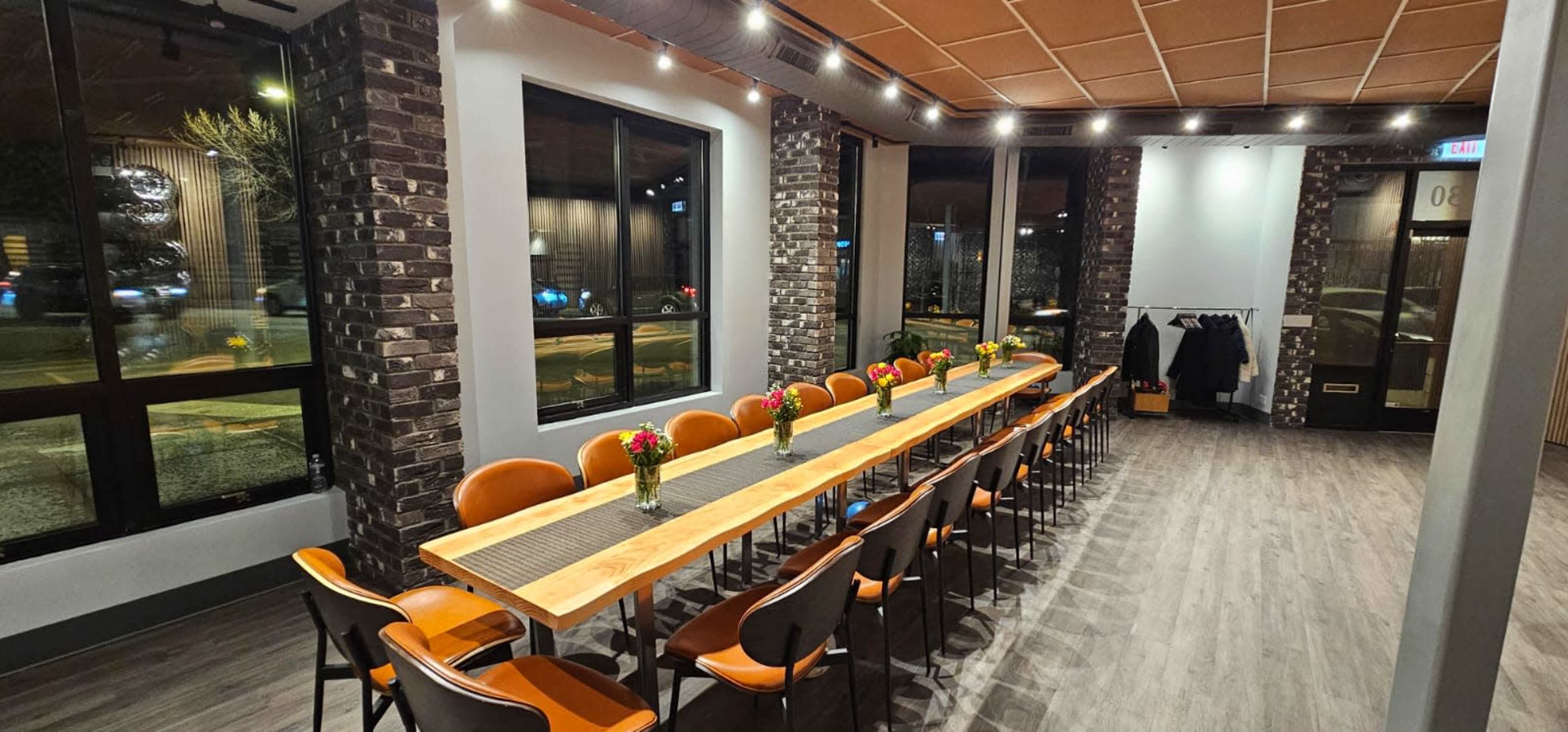 A long wooden dining table set with flower arrangements, surrounded by chairs, in a well-lit restaurant with large windows and brick walls.