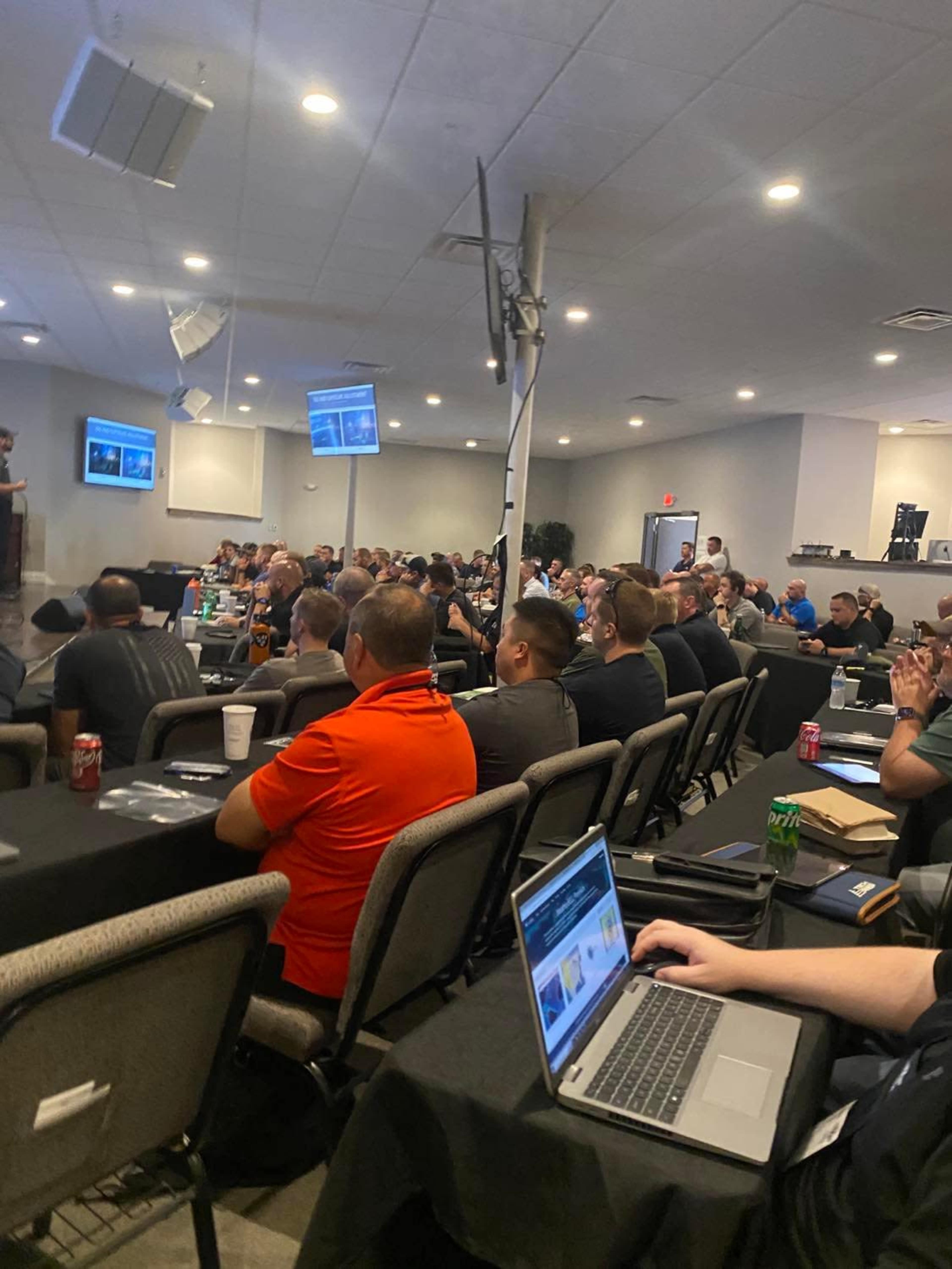 A large group of attendees listens to a presentation in a conference room filled with tables and chairs.