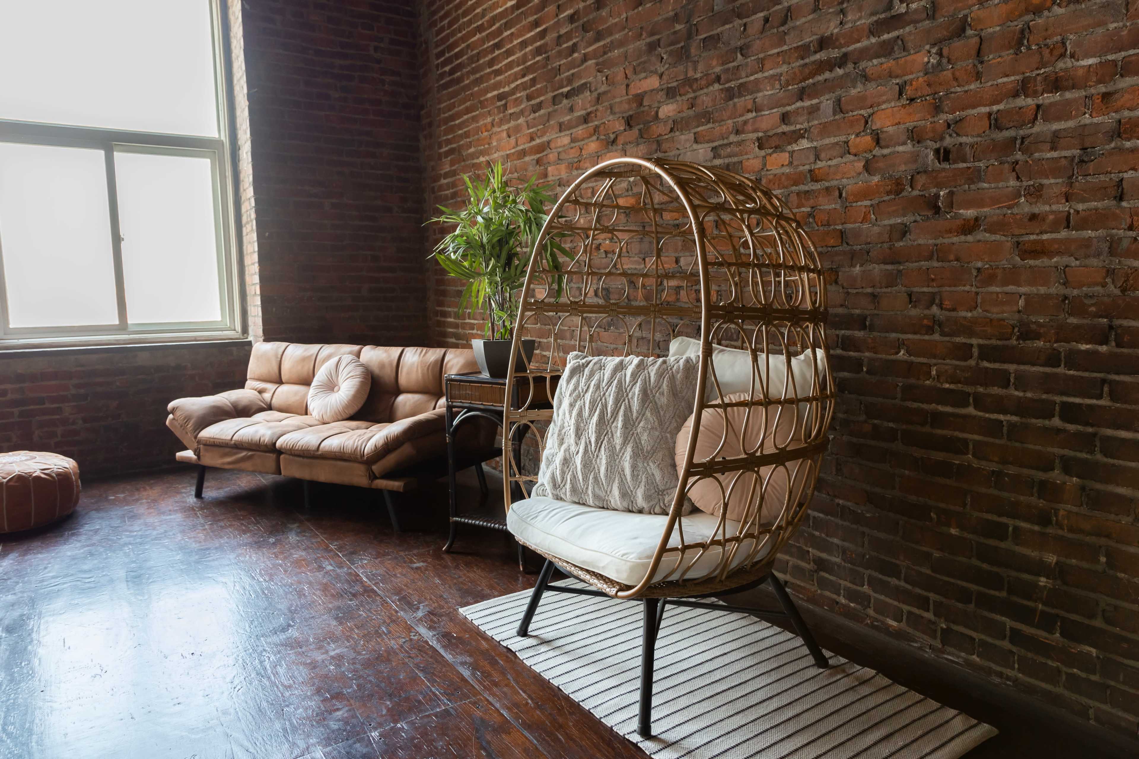 A wicker chair with cushions is positioned next to a leather sofa in a room featuring exposed brick walls and hardwood flooring.