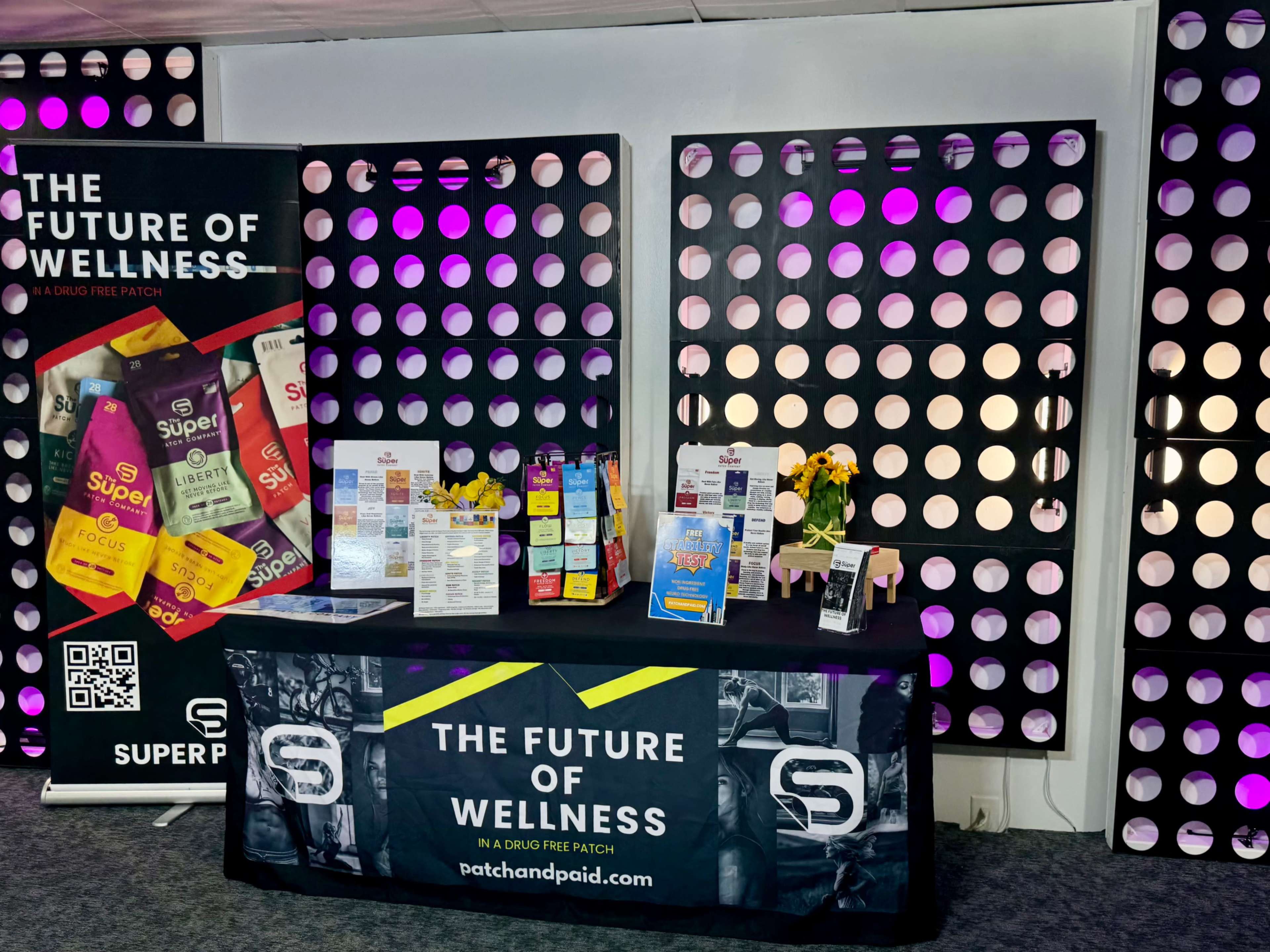 A display booth featuring wellness products and promotional materials is set against a backdrop of purple and black circular patterns.