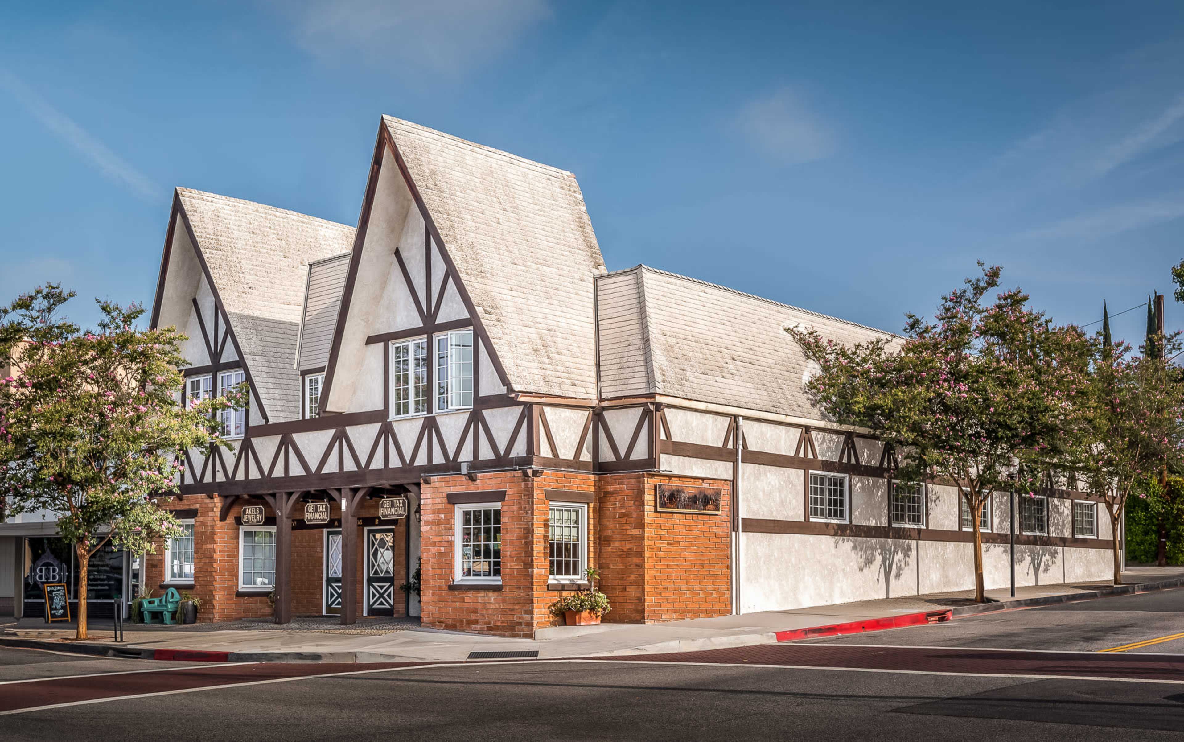 A two-story building with a gabled roof and timber framing is situated on a street corner, surrounded by trees and planters.