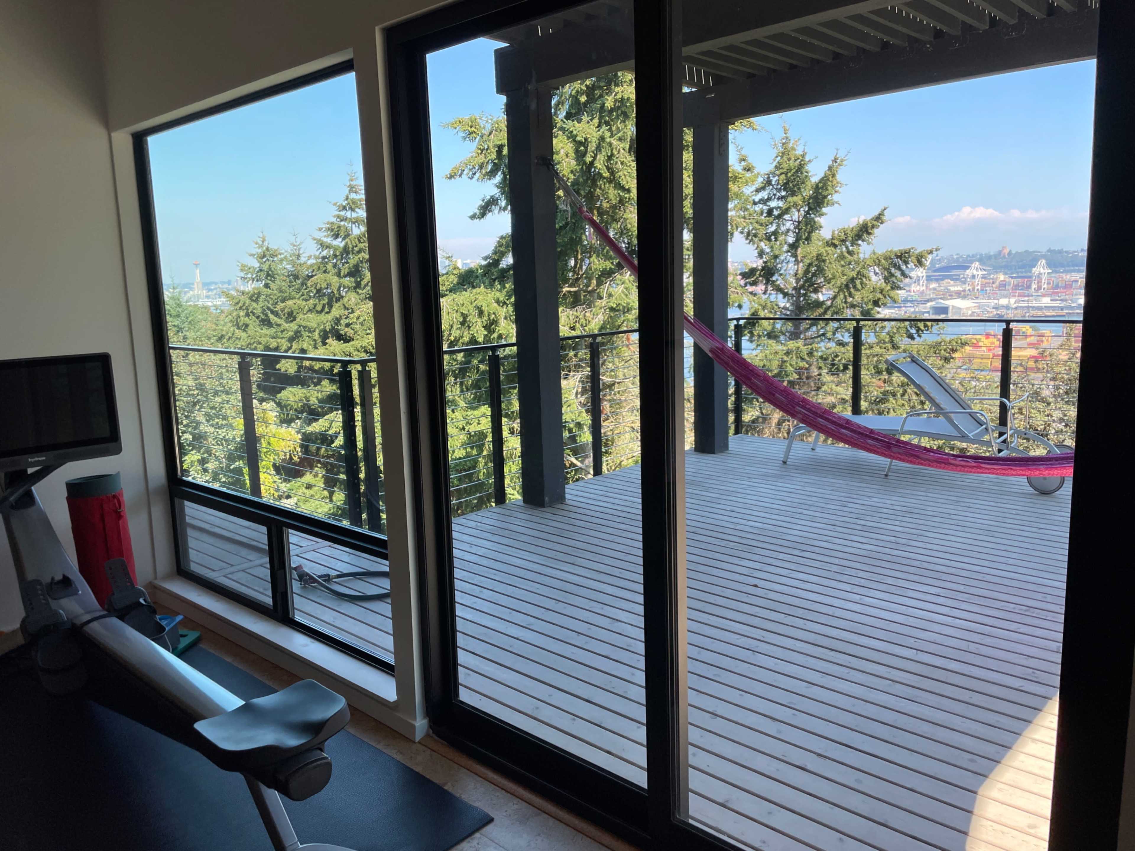 A modern room with large glass doors leading to a wooden deck featuring a hammock, surrounded by trees and a city view in the background.