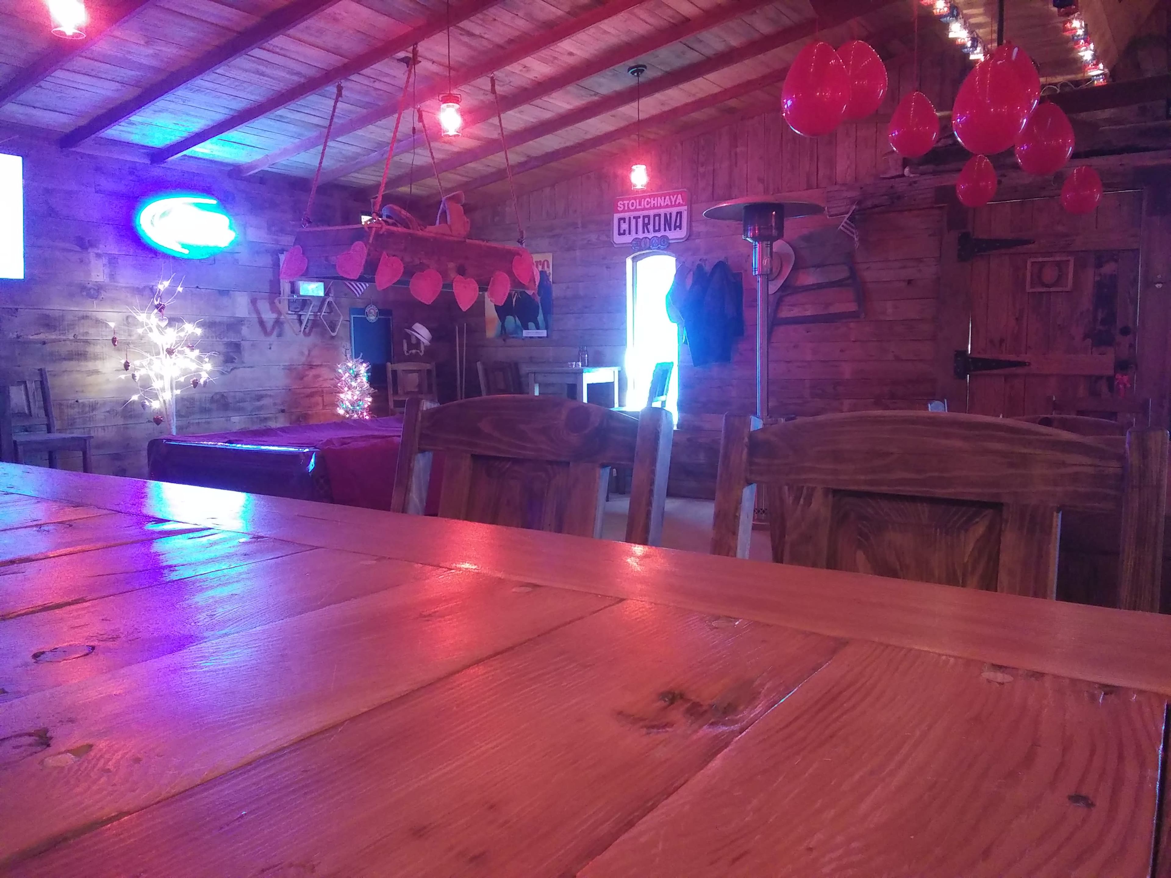 The interior of a rustic wooden cabin features a long table, decorated with red lanterns and dim lighting, with wooden chairs and a door leading outside visible in the background.
