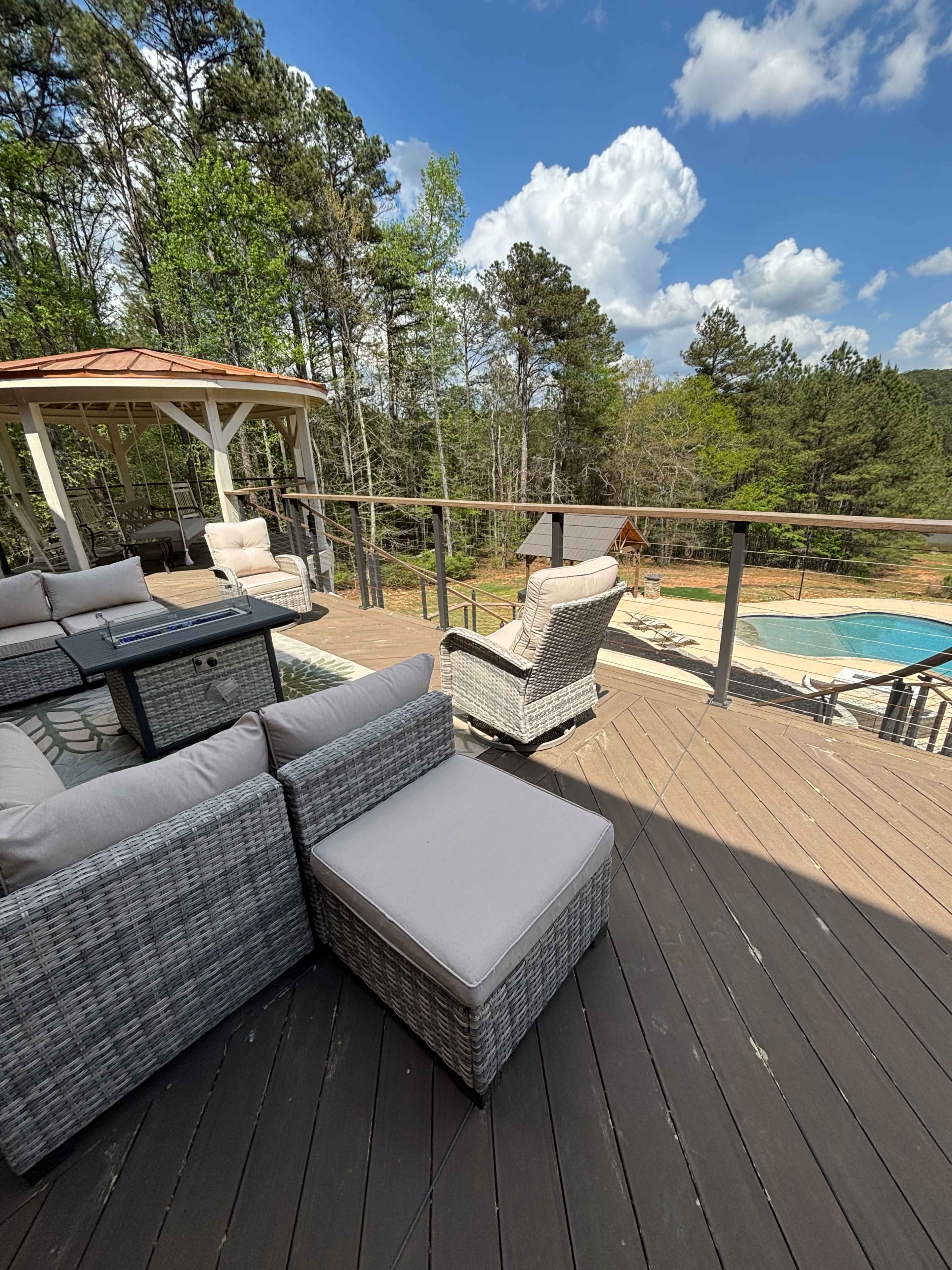 A deck with wicker furniture overlooks a swimming pool surrounded by trees under a partly cloudy sky.