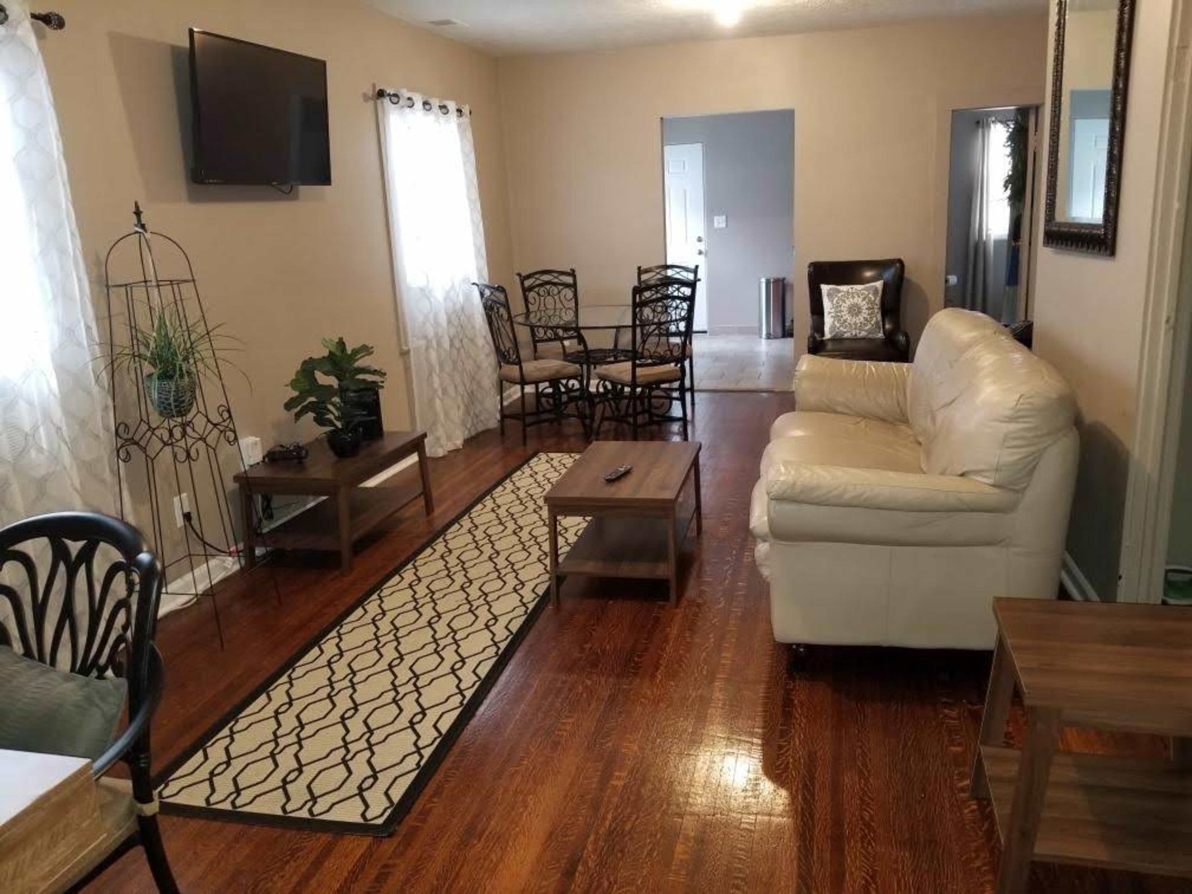 The living room features a white leather sofa, a coffee table, and a dining area with black chairs, all set on polished wood flooring.