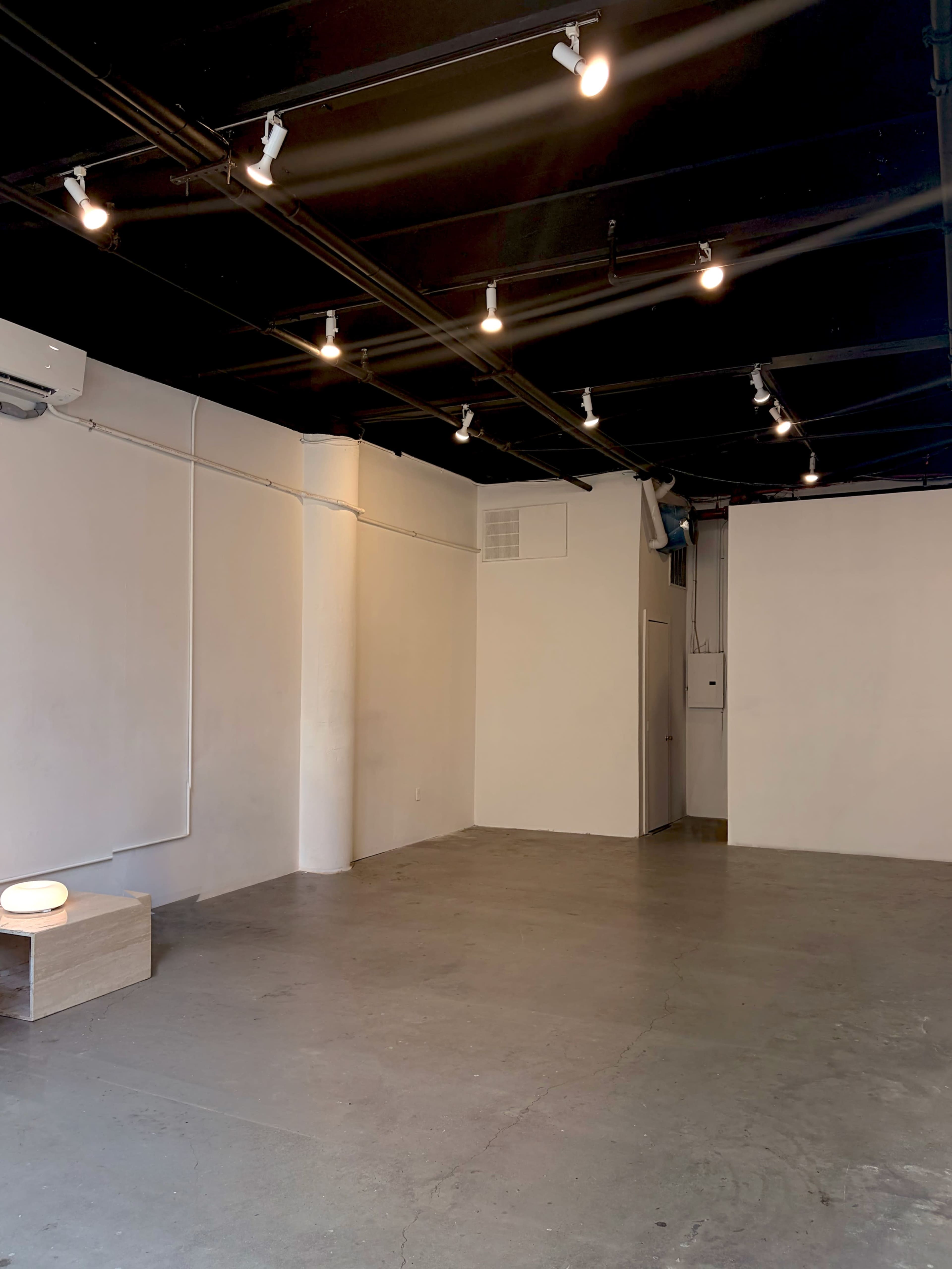 The image shows a sparsely furnished interior space with concrete flooring, white walls, and track lighting along the ceiling.