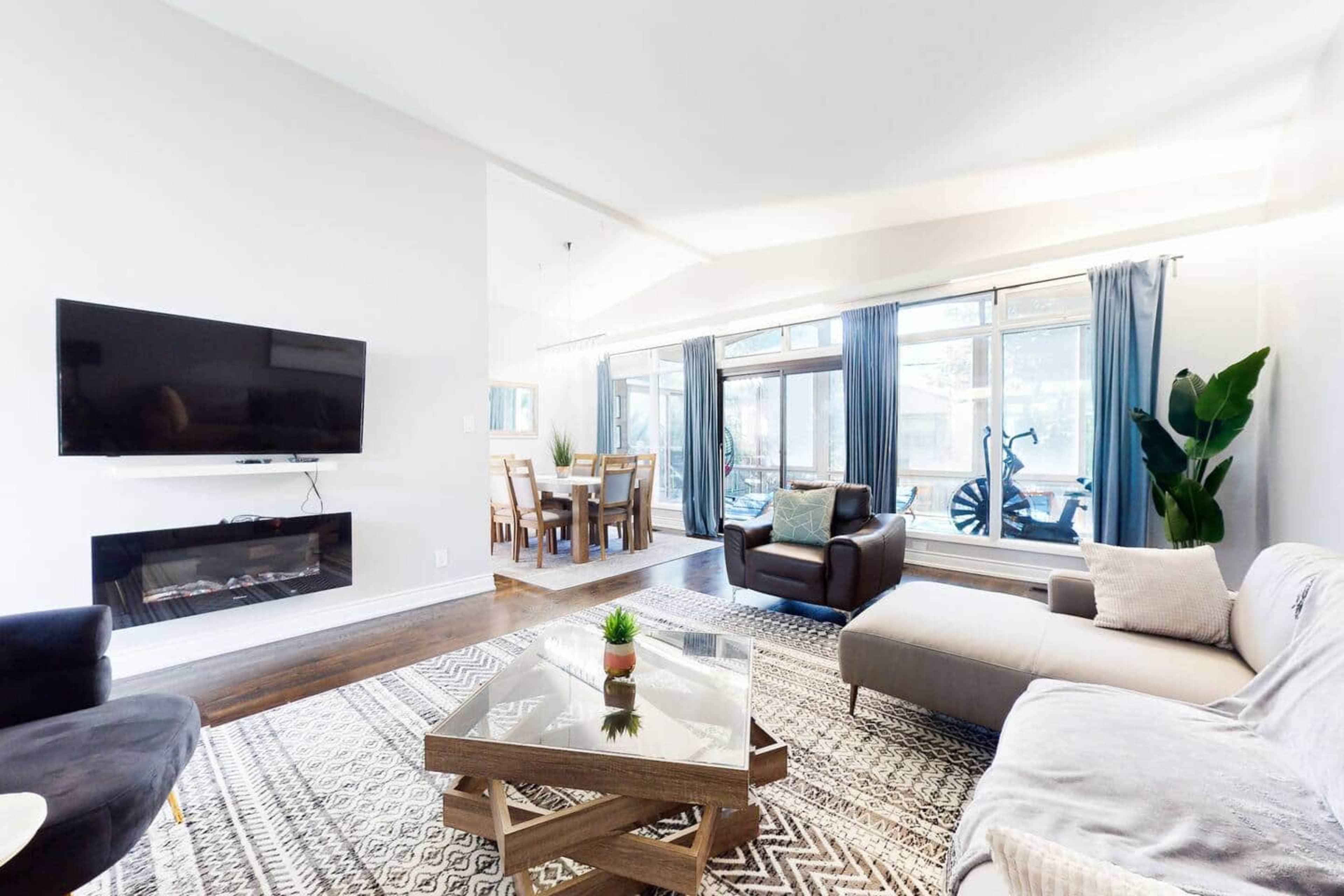 The living room features a modern layout with a flat-screen TV mounted on the wall, a glass coffee table, and large windows allowing natural light to flood the space.