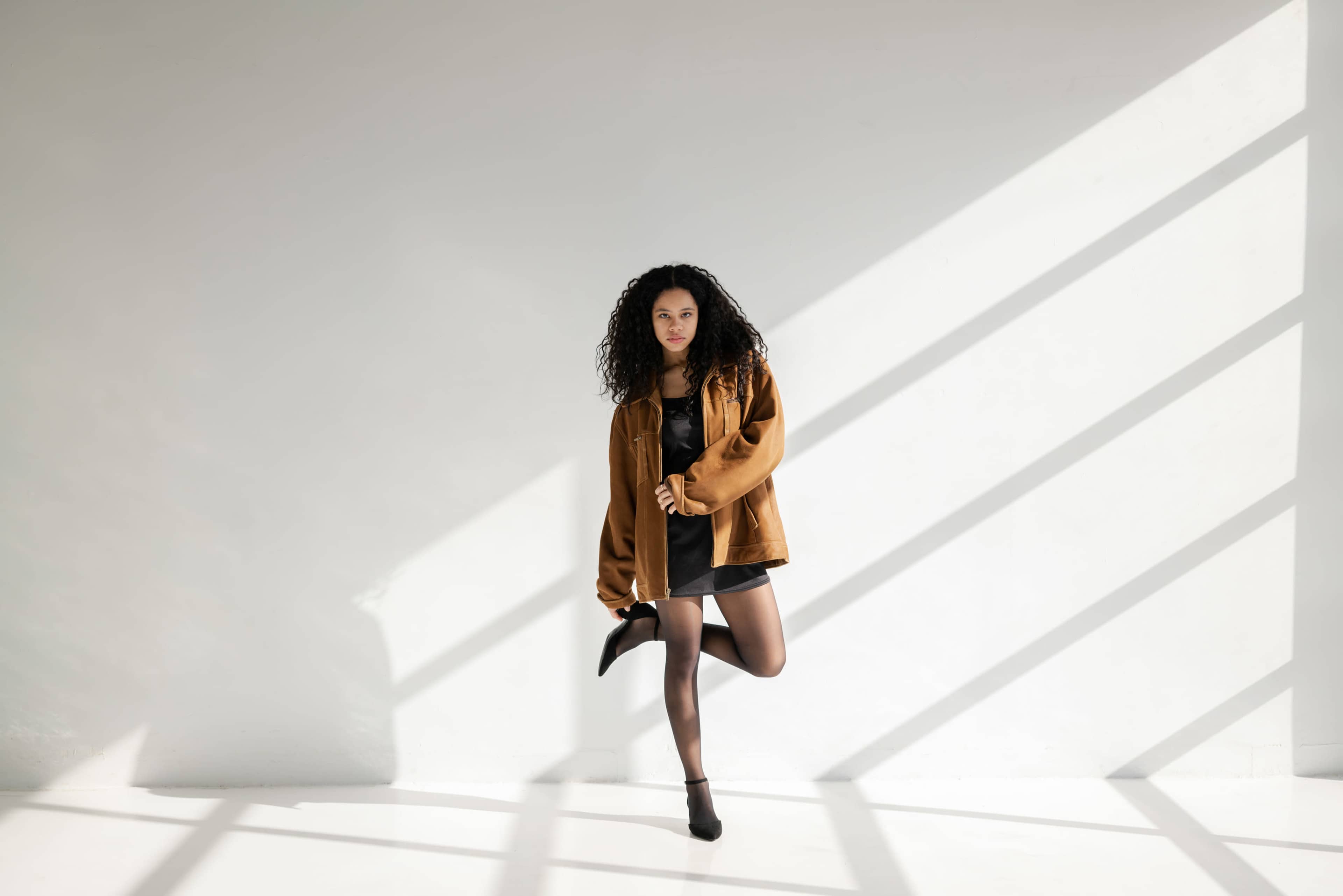 A woman stands in a minimalist space, wearing a brown jacket and black dress, with sunlight casting diagonal shadows on the wall.