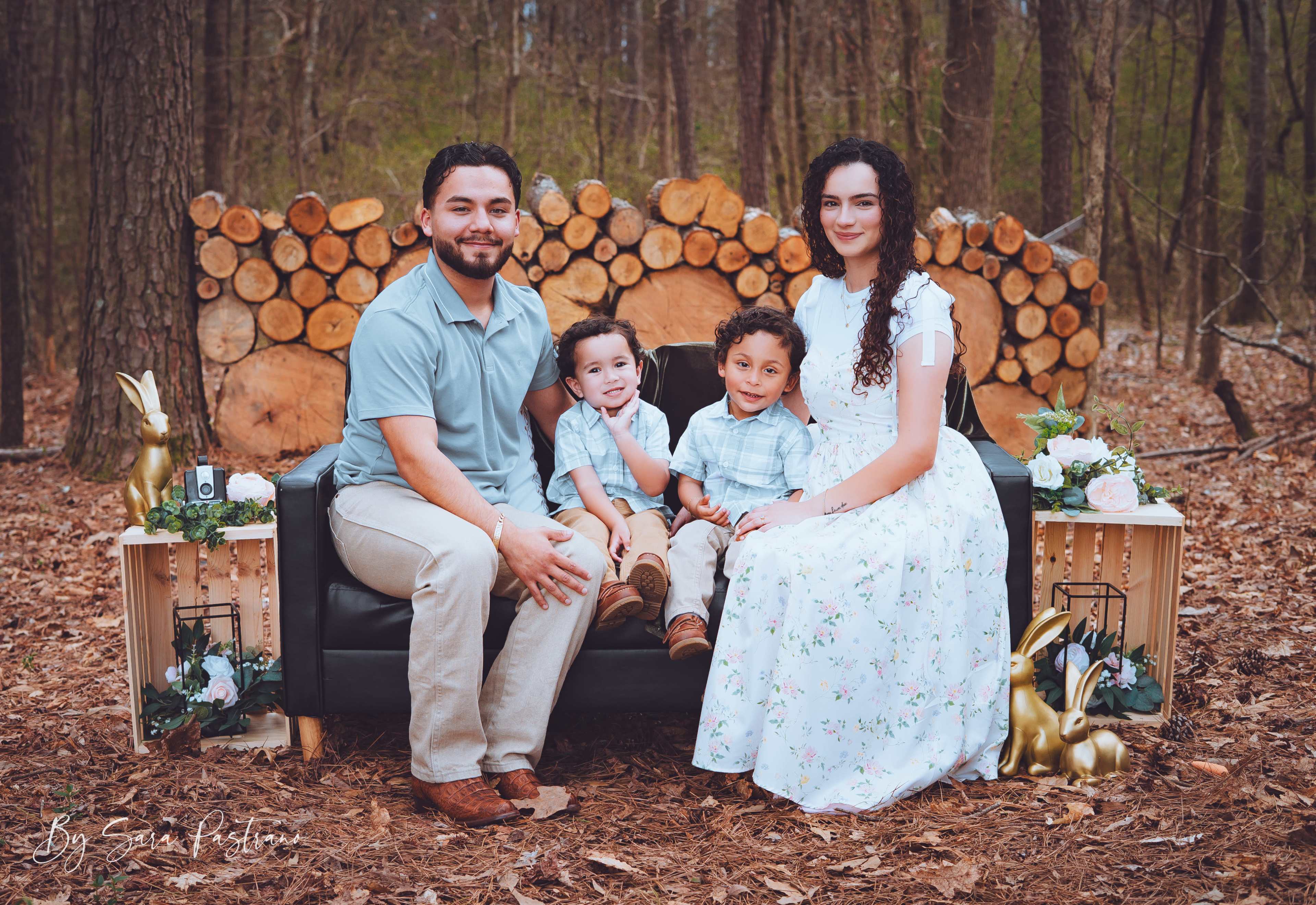 A family of four is seated on a couch in a wooded area, with a backdrop of stacked logs and decorative plants.