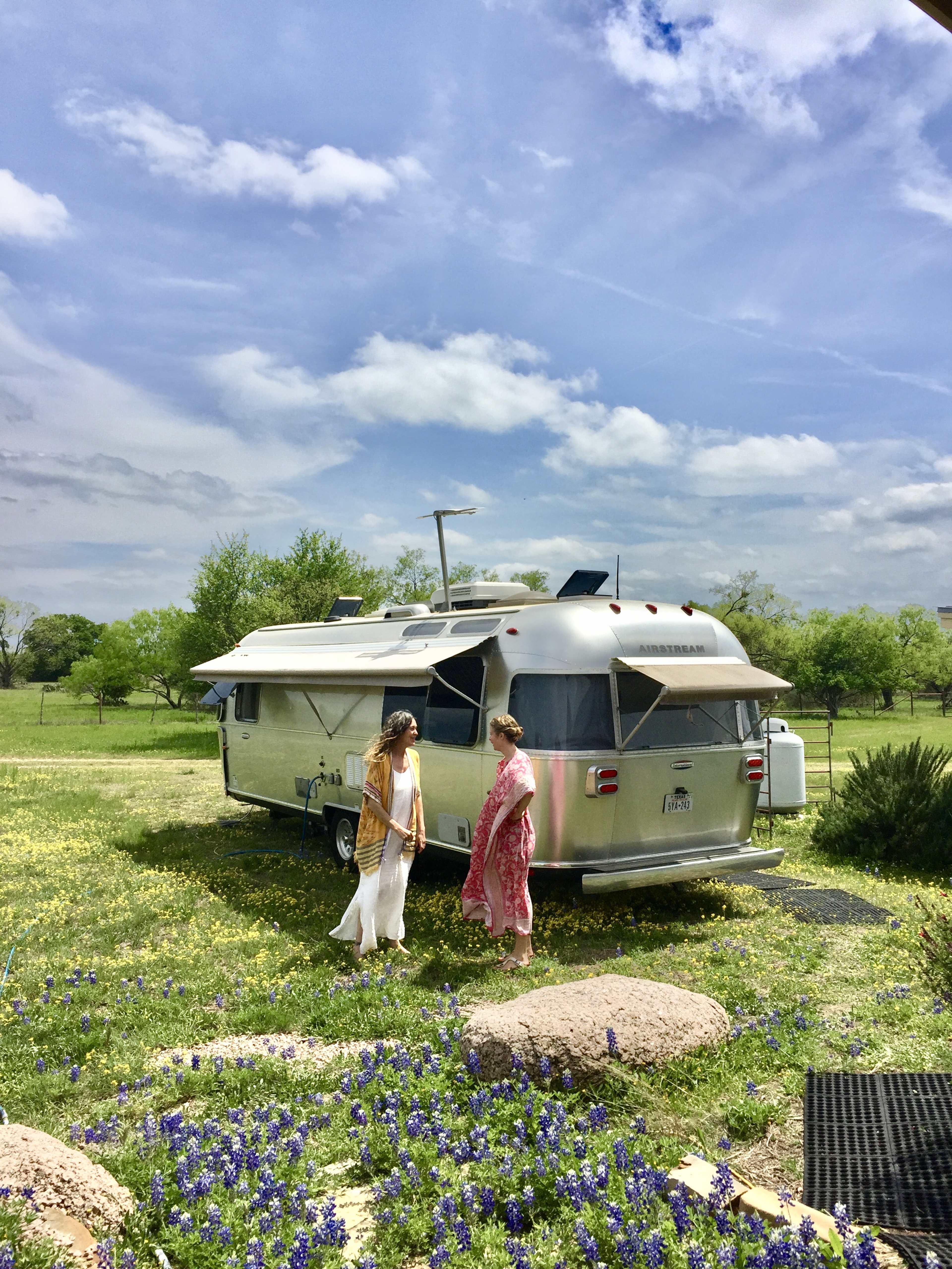 Two women are standing near an Airstream trailer on a grassy field with bluebonnets and trees in the background.