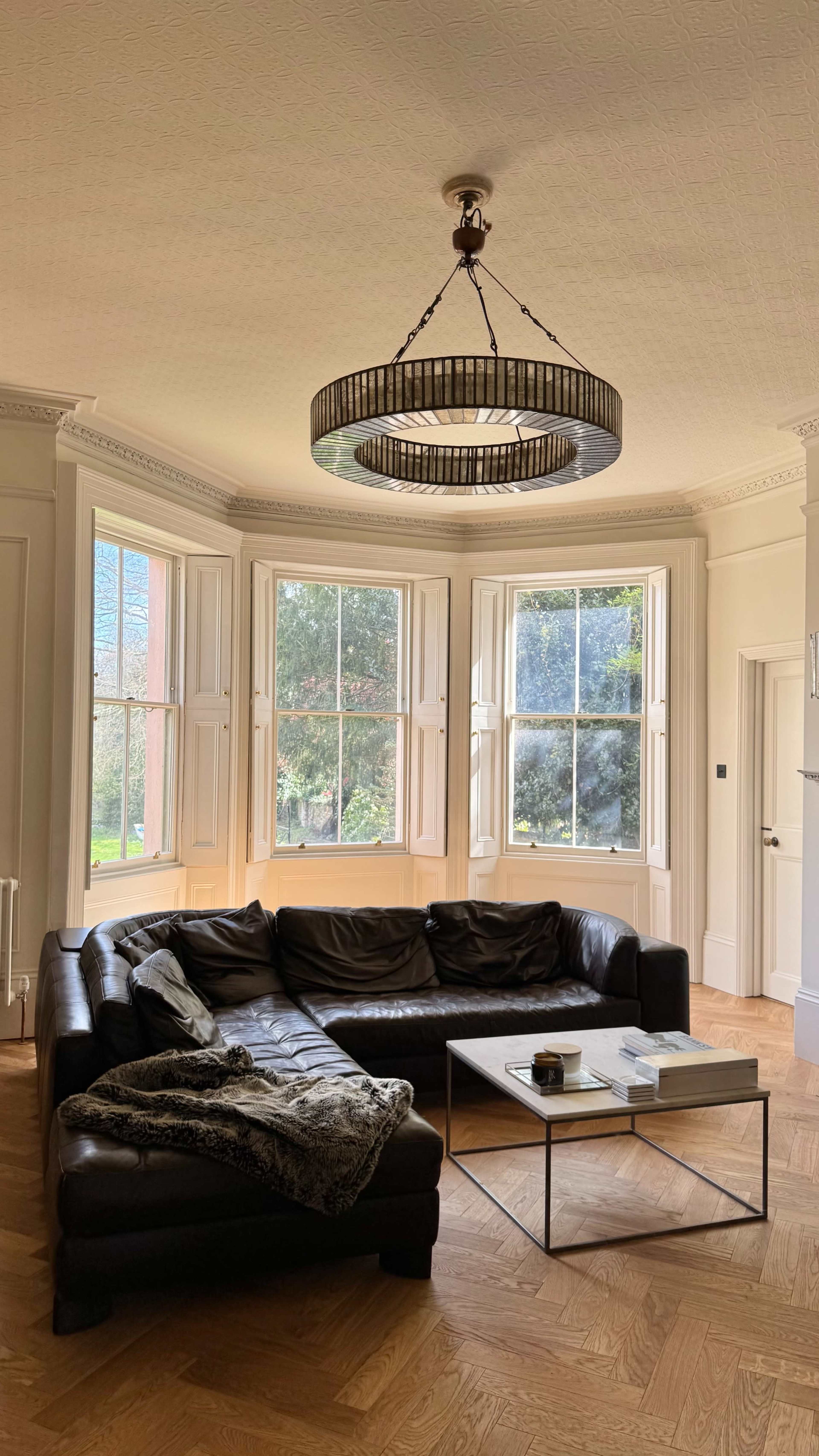 The image shows a spacious living room with a large black leather couch, a coffee table in front of it, and three large windows allowing natural light to enter the space.