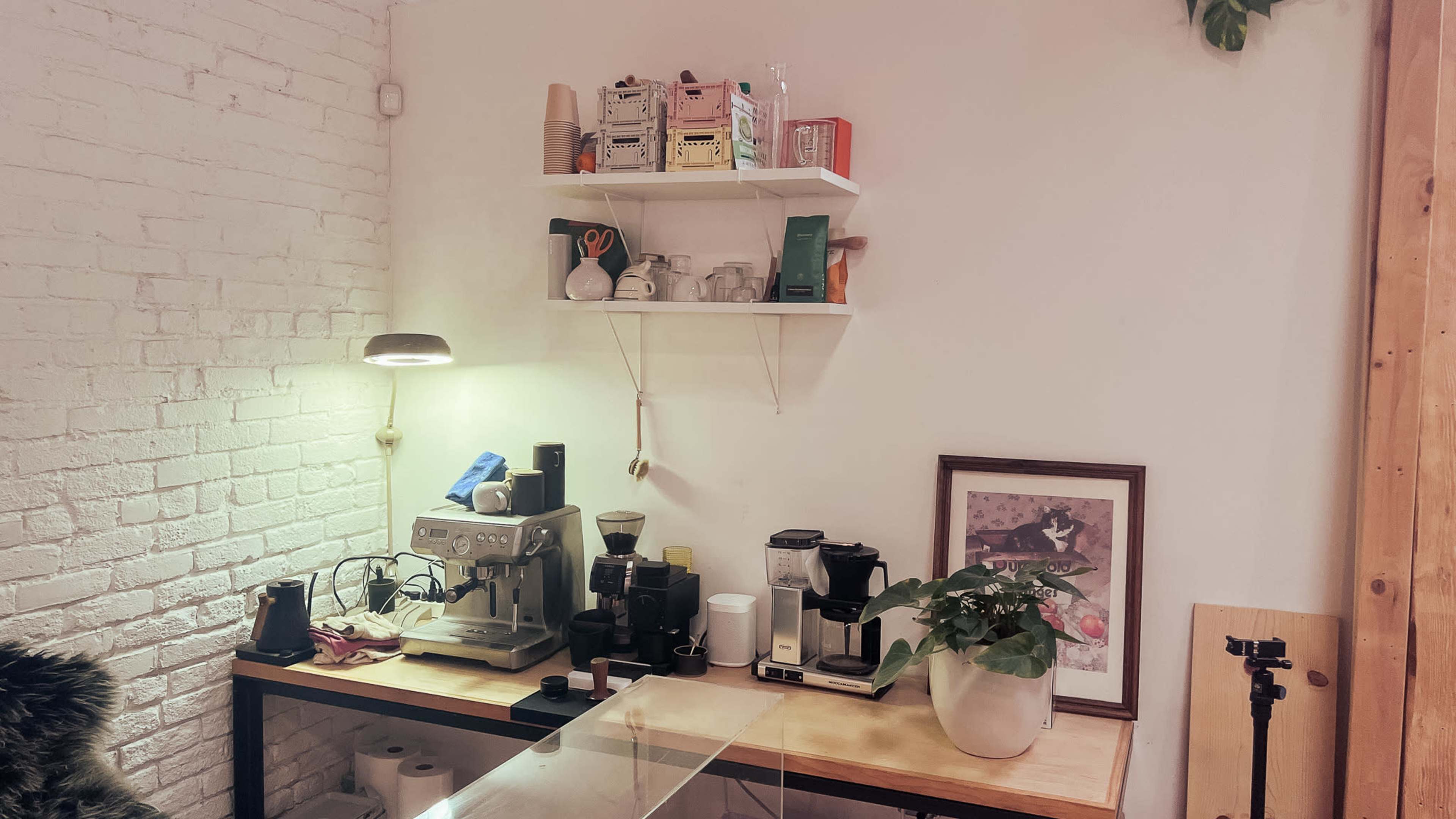 A small workspace features a coffee machine setup, shelves with various coffee supplies, a plant, and framed artwork against a white brick wall.