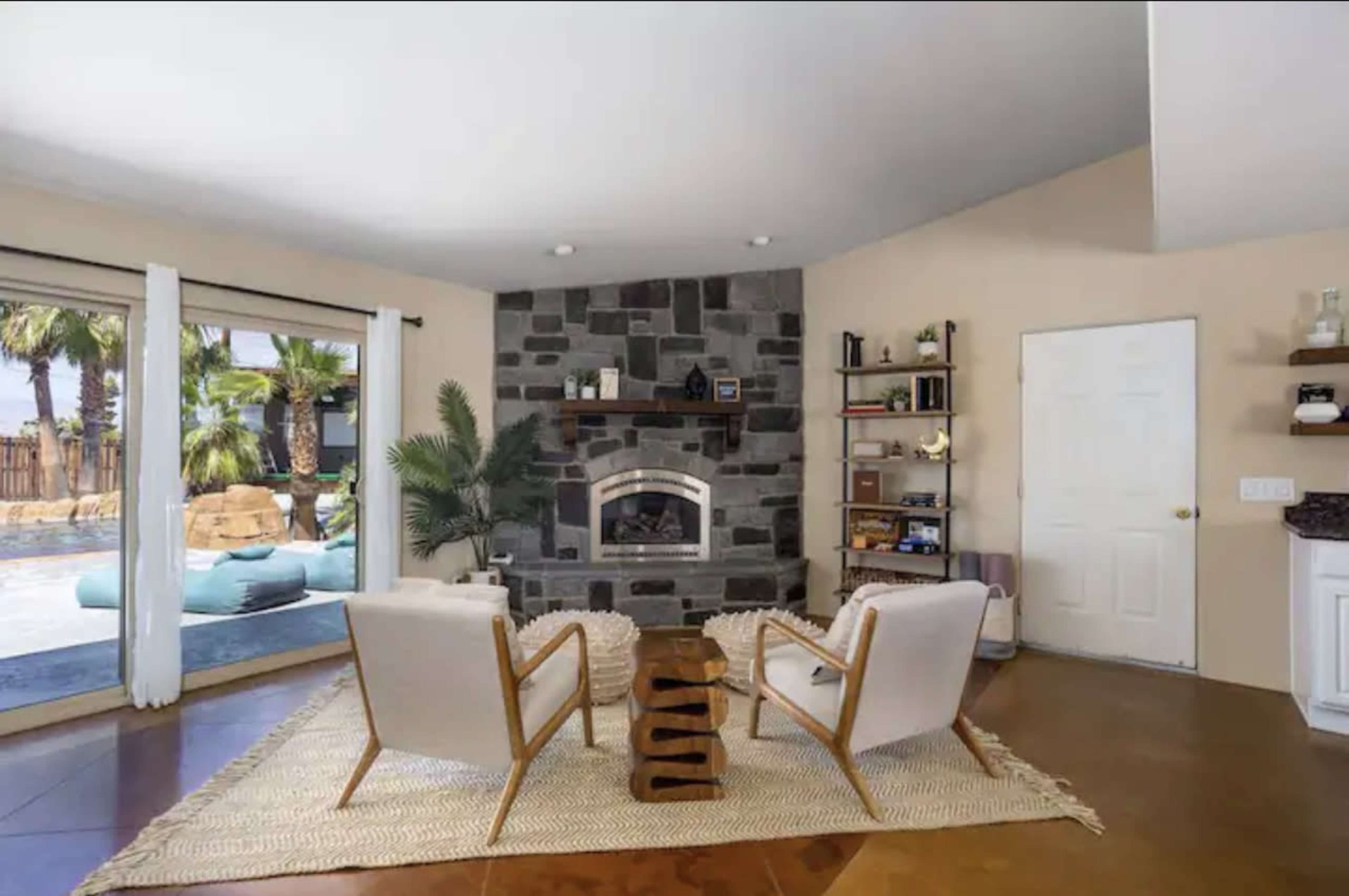 A modern living room features two chairs facing a stone fireplace, with a sliding glass door leading to an outdoor pool area.