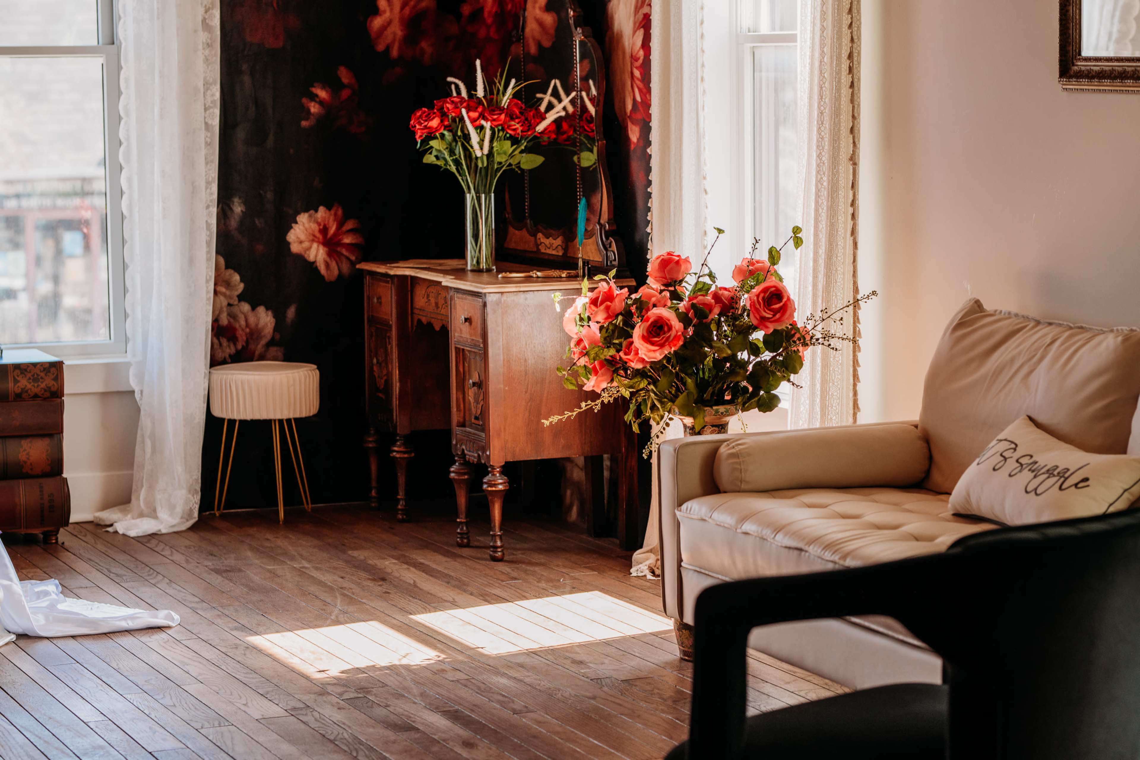 The image shows a cozy interior featuring a beige sofa, a small decorative table, a wooden sideboard, and flower arrangements placed in a well-lit room with floral wallpaper.