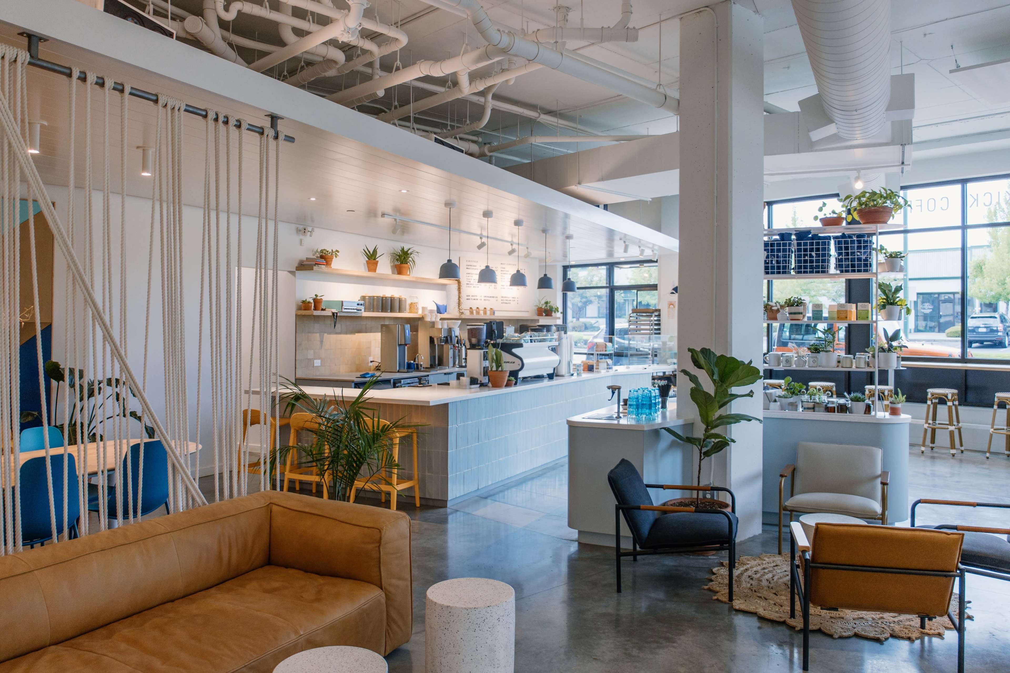 A modern café interior with a light color scheme, featuring seating areas, a coffee bar, and plants.