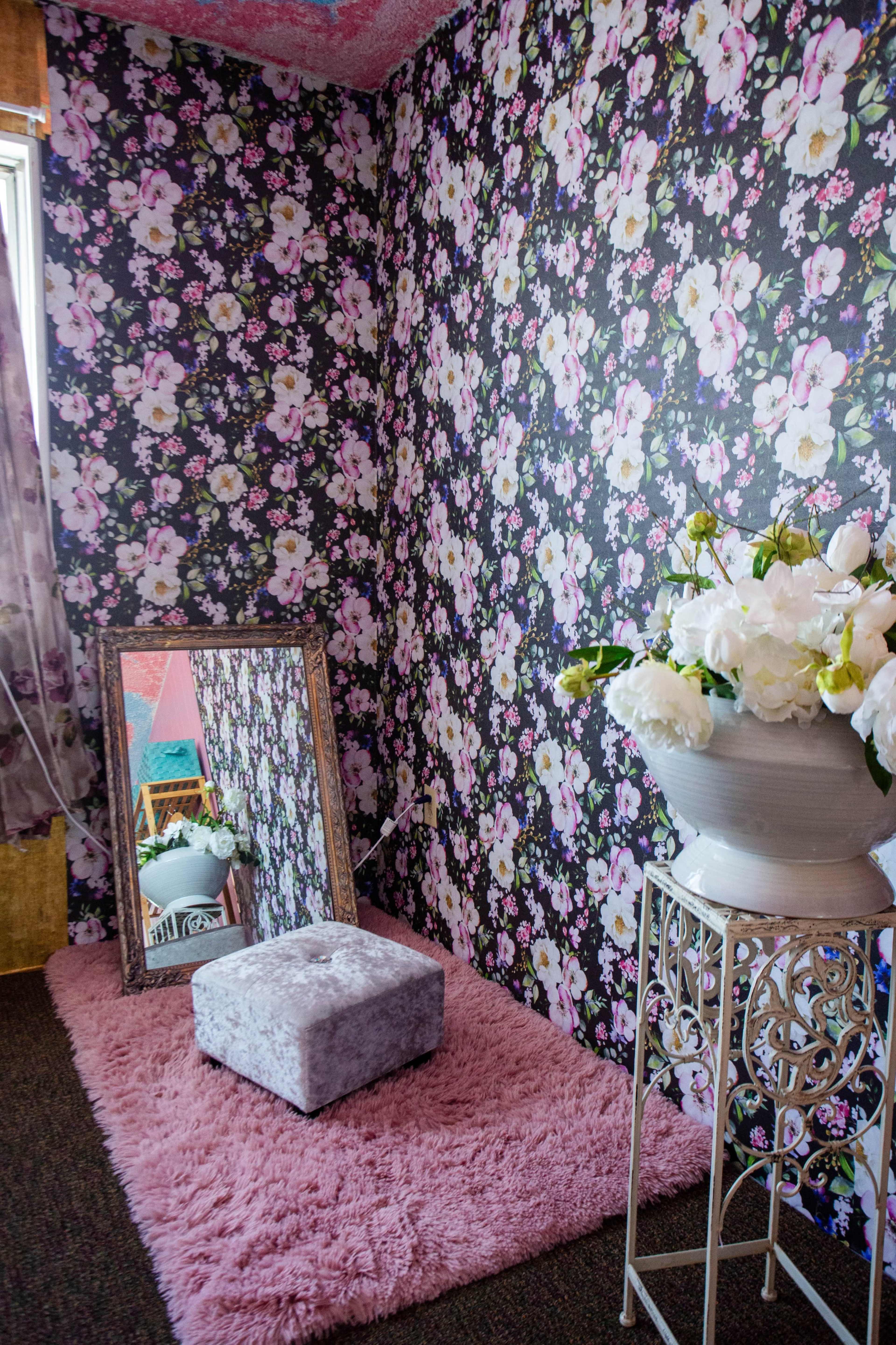 A corner of a room features floral wallpaper, a large mirror, a small gray ottoman, a fluffy pink rug, and a vase with white flowers on a decorative stand.