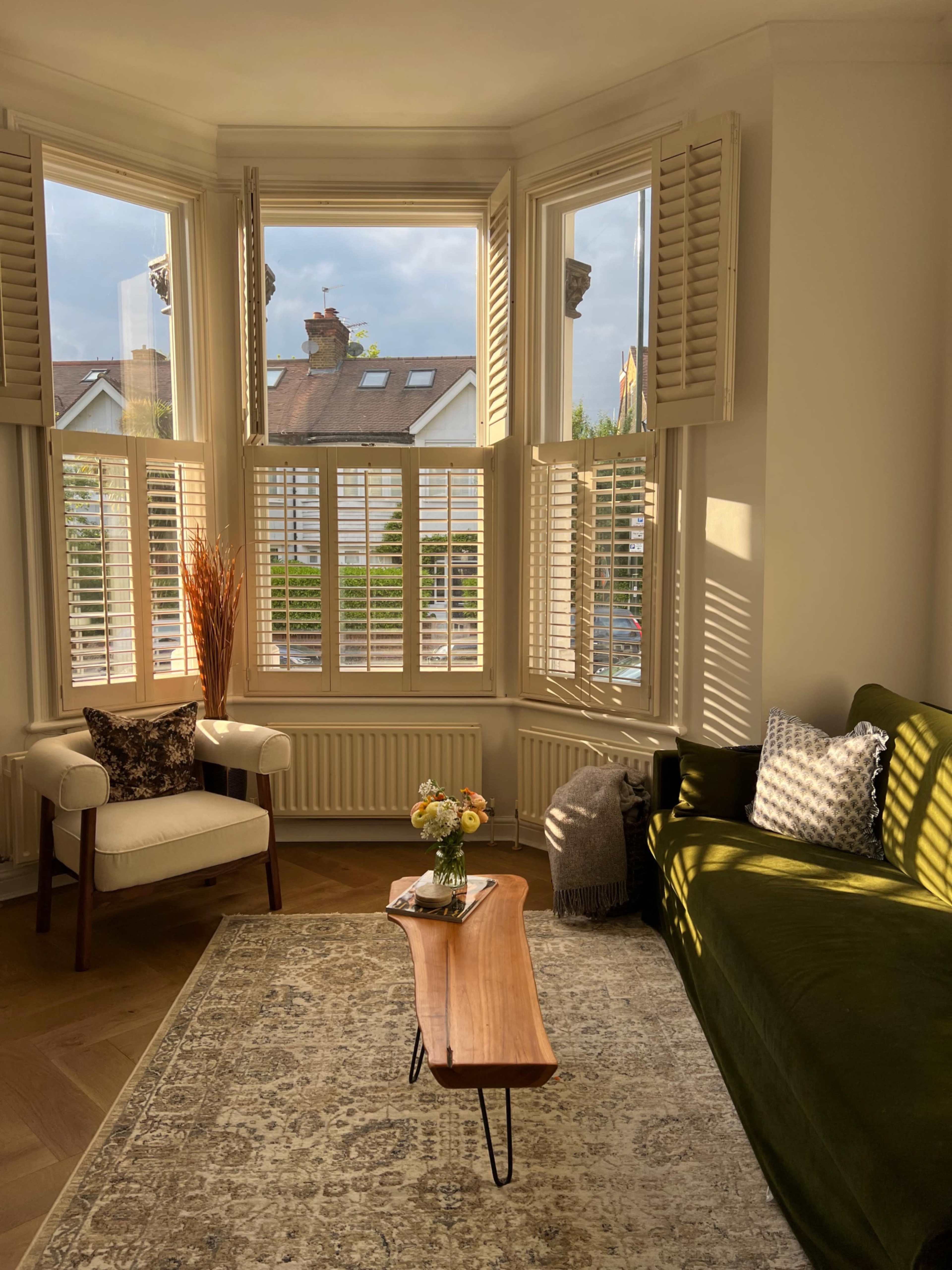 A cozy living room features a green sofa, a small wooden coffee table, and a comfortable armchair near large bay windows with shutters.
