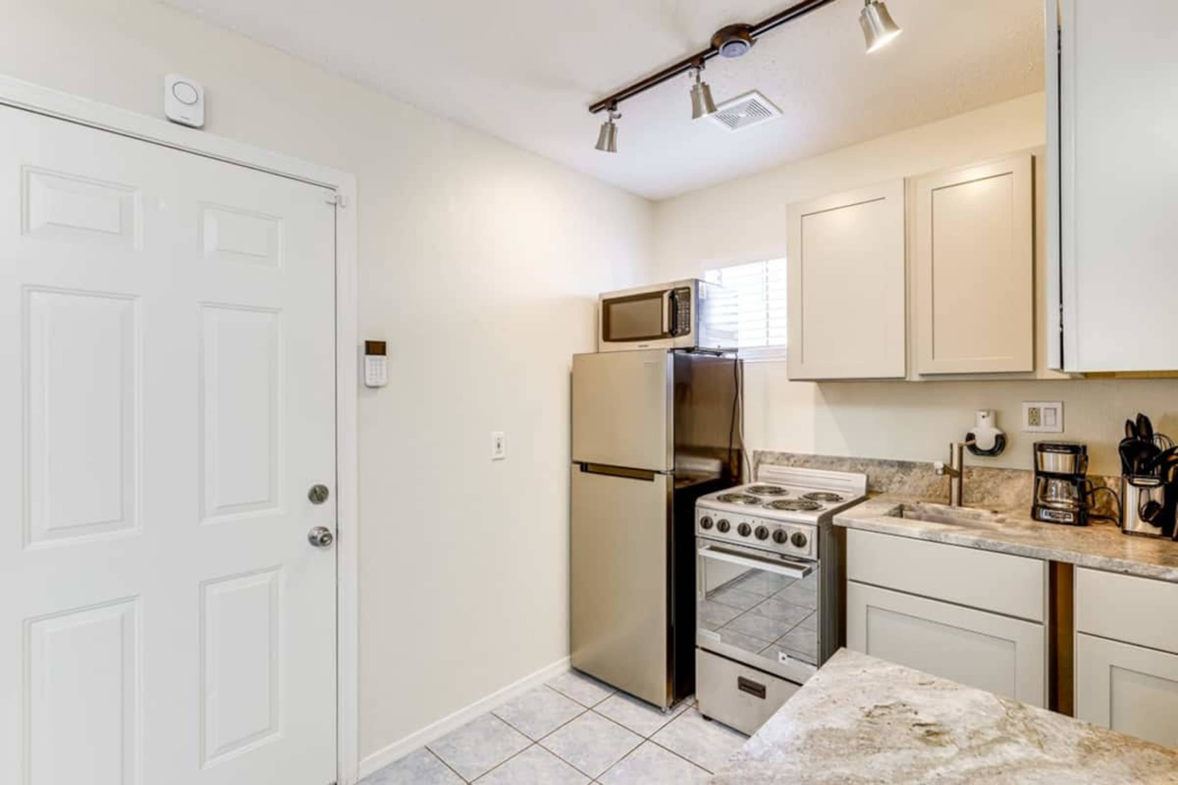 The image shows a compact kitchen with a stainless steel refrigerator, an electric stove, a microwave on top, and light-colored cabinets and countertops.
