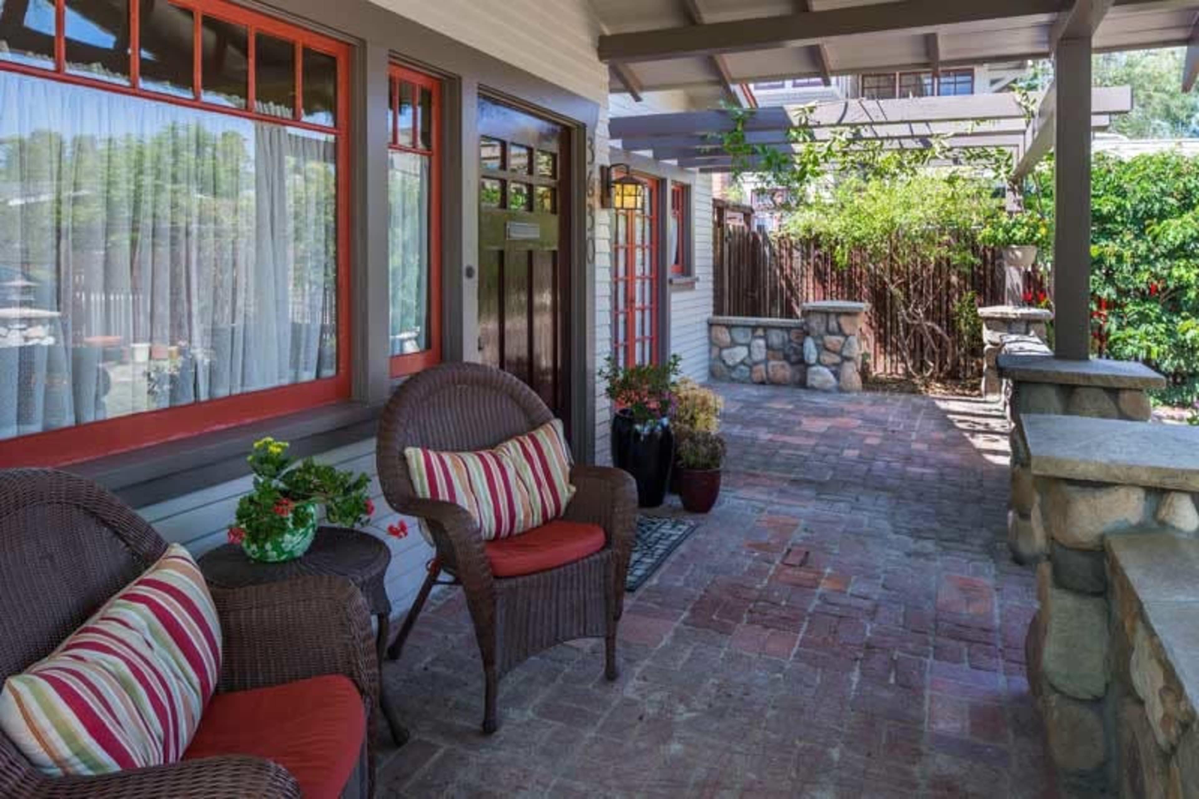The image shows a porch with two wicker chairs adorned with striped cushions, brick flooring, and potted plants against a backdrop of a wooden door and a garden.