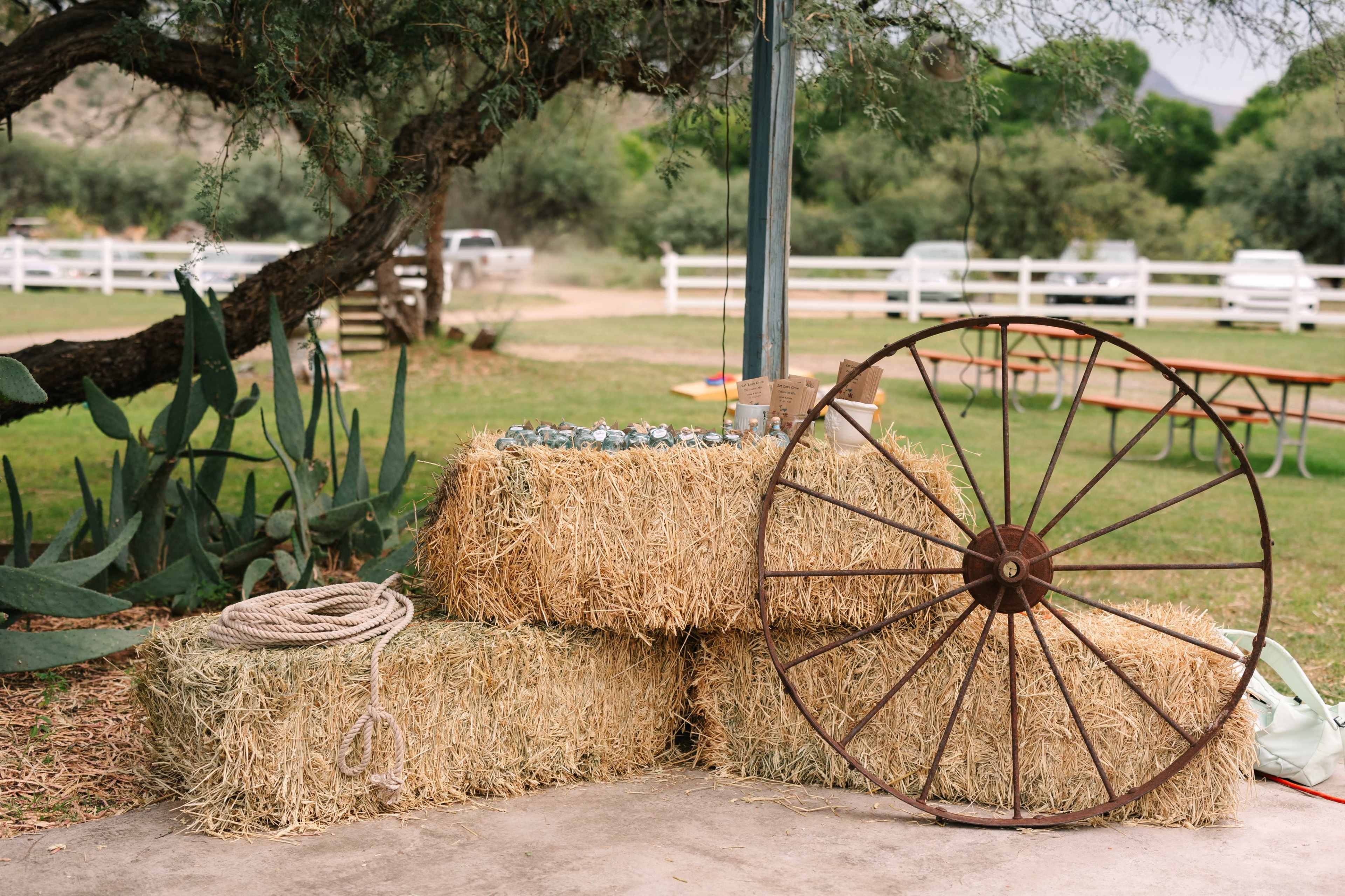 A stack of hay bales is placed beside a wooden wagon wheel in a grassy outdoor setting.