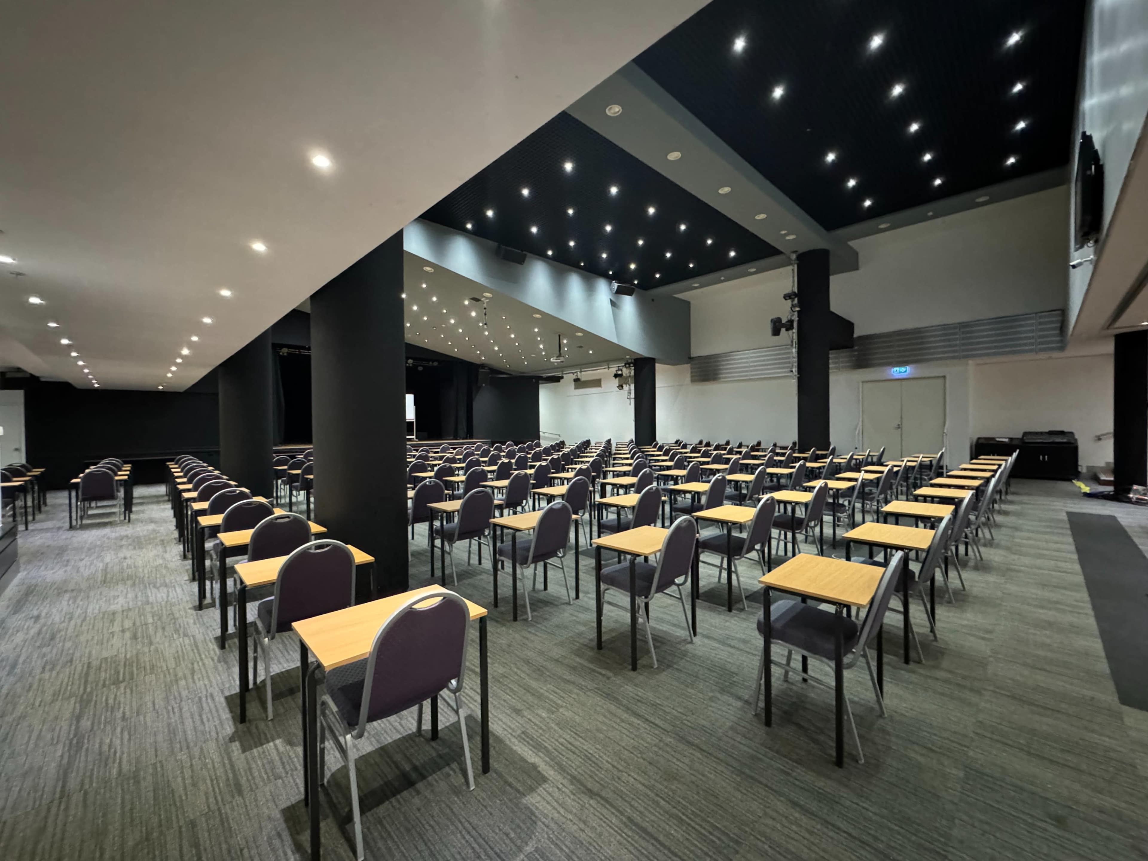 The image shows a large conference room filled with rows of empty tables and chairs under a ceiling with numerous lights.